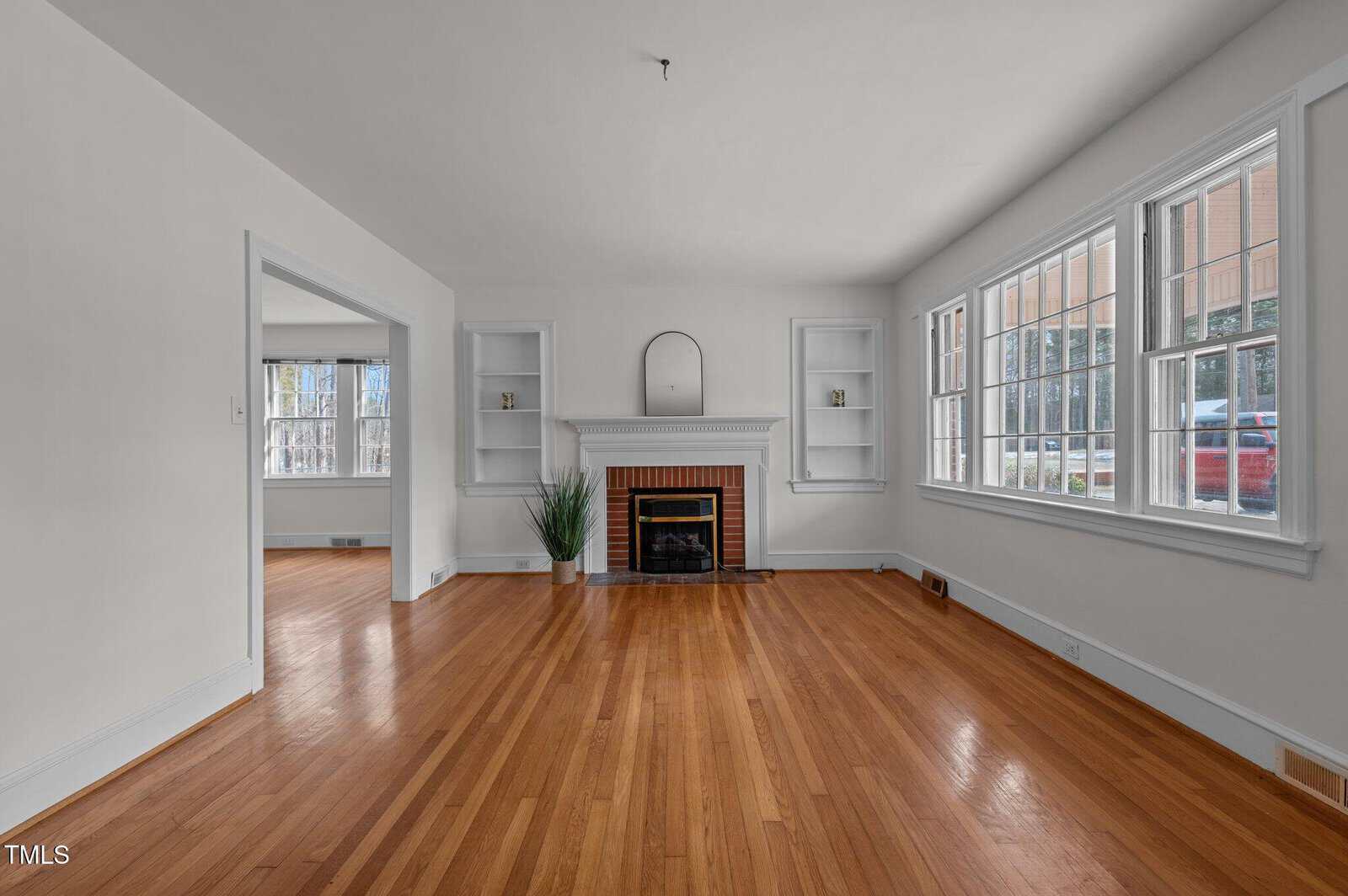 An empty living room with wooden floors, white walls, and a white fireplace with a small mirror above it. Two built-in shelves flank the fireplace. Large windows on the right let in natural light. A small potted plant sits on the floor near the fireplace.
