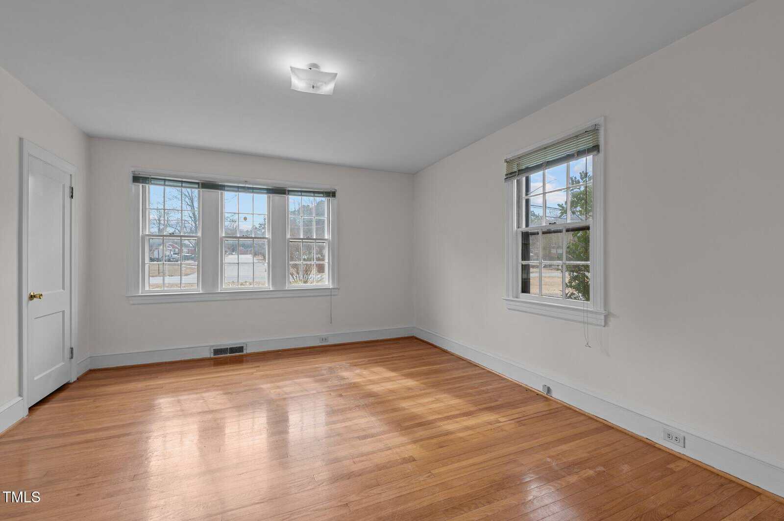 Empty room with hardwood floors, cream-colored walls, two large windows on one wall, a smaller window on another wall, a closed white door, and a ceiling light fixture. Sunlight is streaming in through the windows.