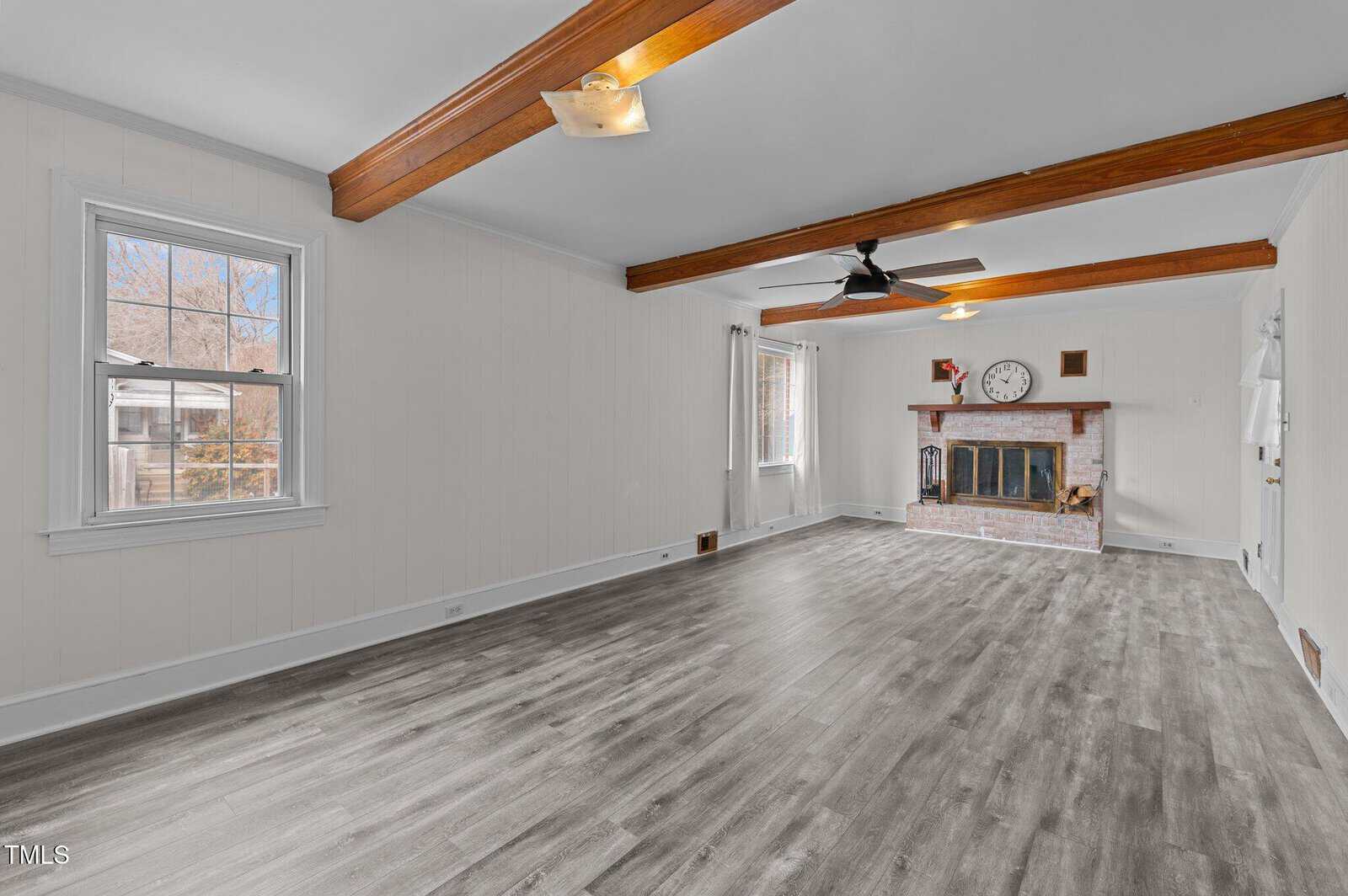 A spacious living room with light wood flooring, white walls, and wooden ceiling beams. A brick fireplace with a clock and decor sits at one end. Two windows and a glass door allow natural light to enter. A ceiling fan is in the center.