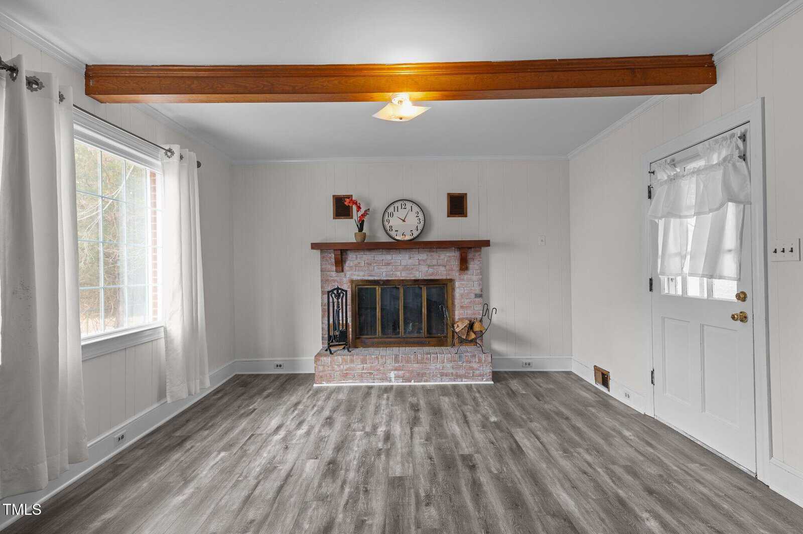 A living room featuring a brick fireplace with a clock and small statue on the mantel. The room has light-colored walls, a ceiling beam, large window with curtains, and a glass-paneled door. The floor is gray wood.