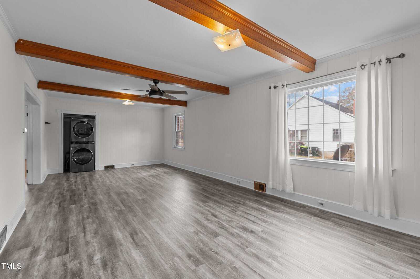 A bright room with light gray laminate flooring, white walls, and wooden ceiling beams. A ceiling fan hangs centrally. There's a window with white curtains on the right, and a stacked washer and dryer are visible in the back.