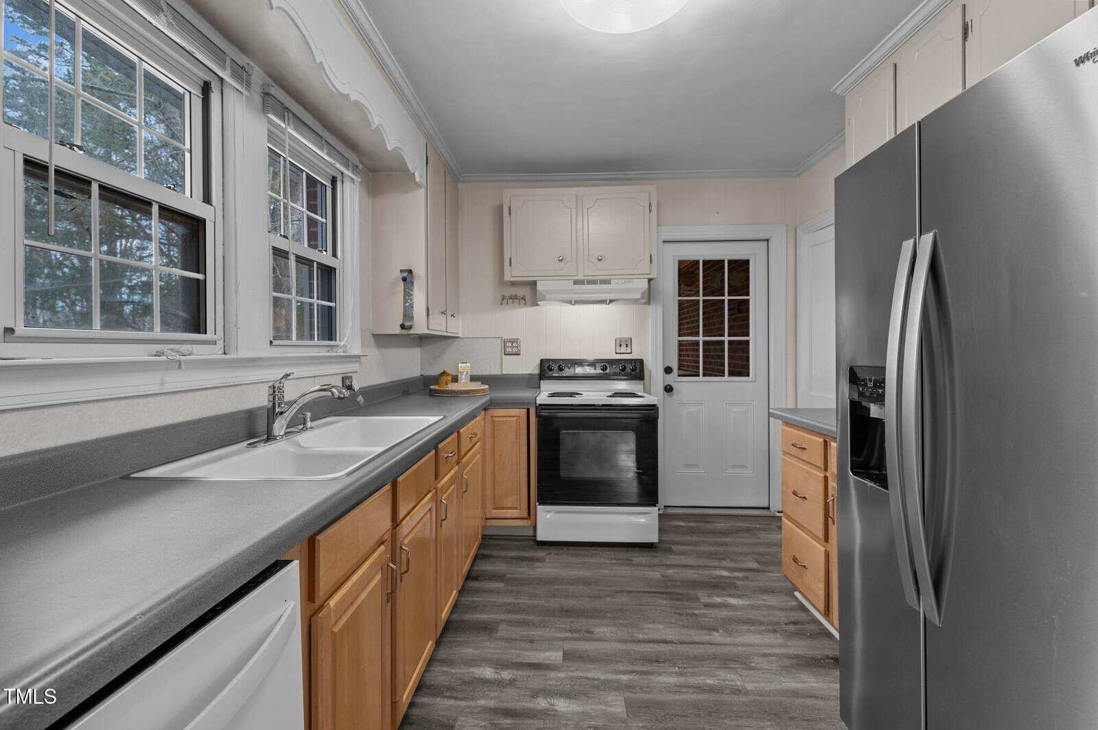 A kitchen featuring wooden cabinets, a light gray countertop, and a stainless steel refrigerator. It includes a double sink, an electric stove with an oven, and a window above the sink. The floor is made of dark wood, and there is a door with glass panels.
