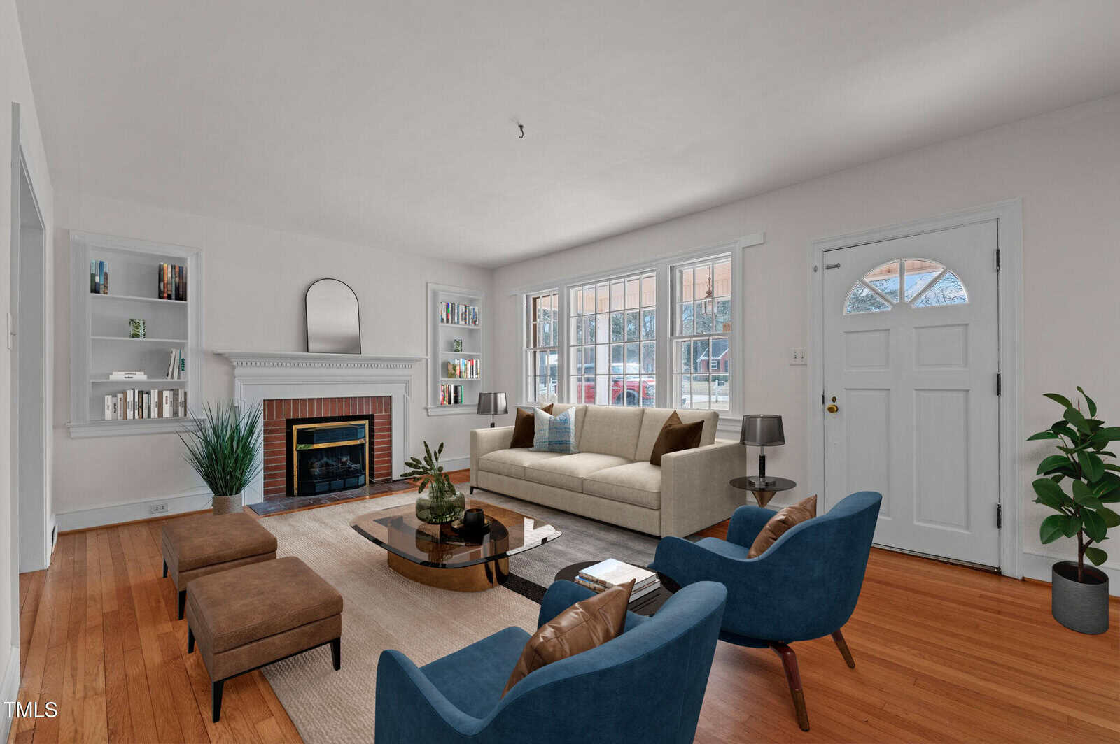A living room with white walls and hardwood floors features a beige sofa, two blue chairs, a glass-top coffee table, and two brown ottomans. A fireplace with a wood mantel is on the left, and large windows and a white front door are on the right.