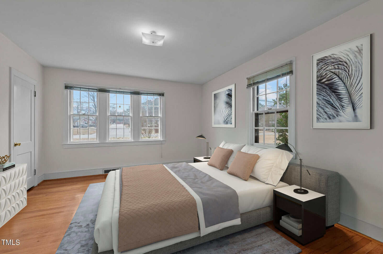 A bedroom with a large bed, two nightstands, lamps, framed artwork on light walls, three windows letting in daylight, and a hardwood floor with a rug.