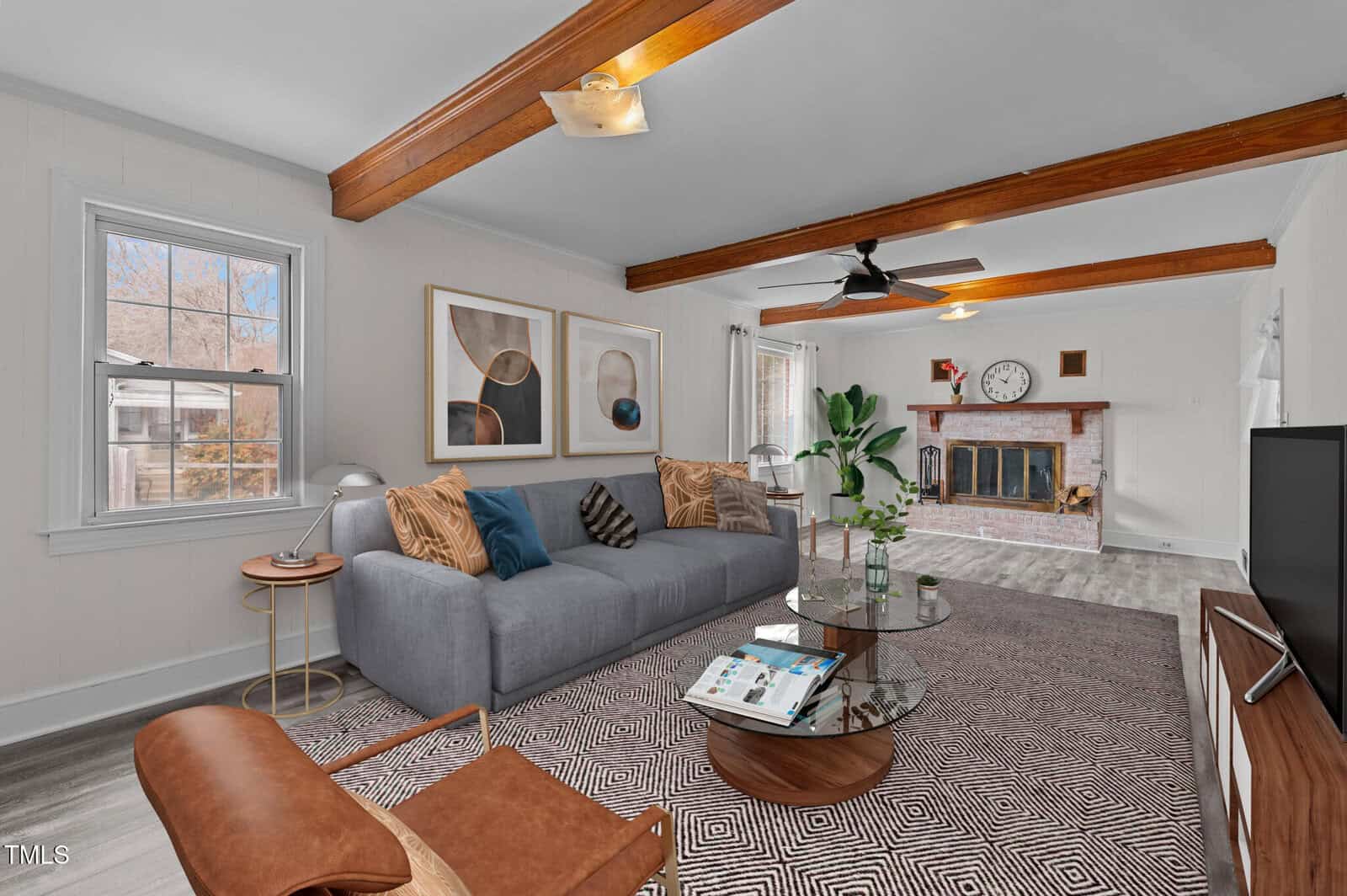 A living room with a gray sofa, adorned with pillows, facing a glass coffee table and a TV. A fireplace is on the far wall with two abstract art pieces above the sofa. Wooden ceiling beams and a patterned rug add warmth to the space.