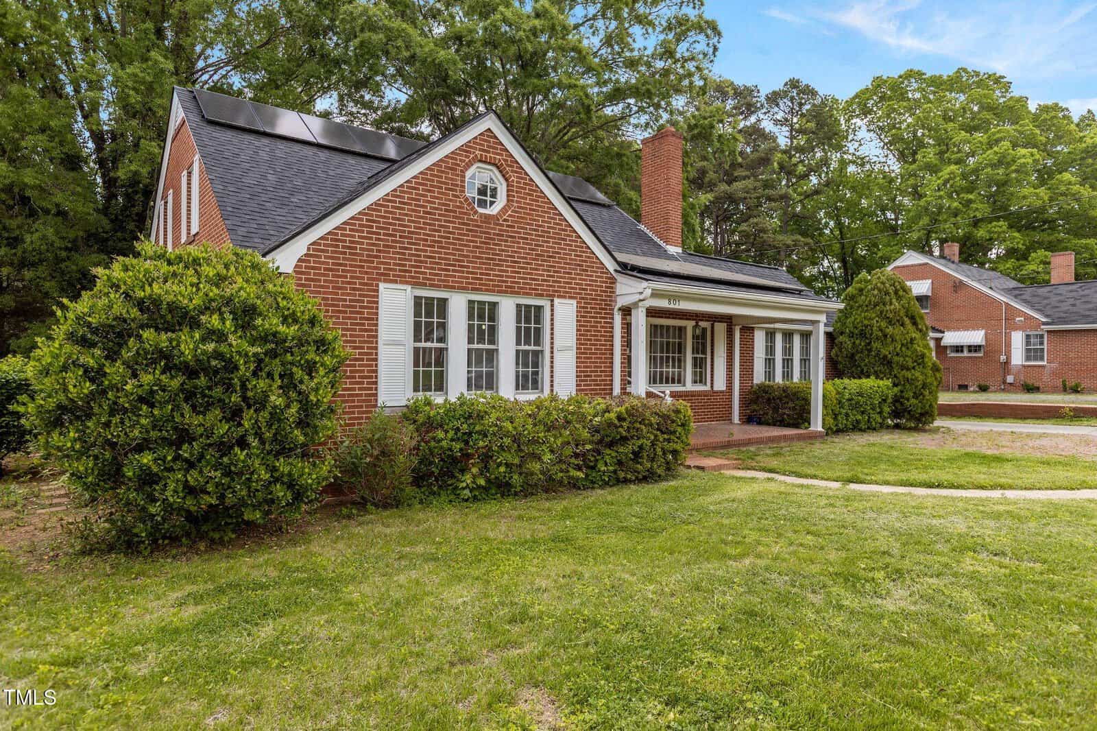 A red brick house with white trim and shutters, solar panels on the roof, a covered porch, and green bushes in front, set on a grassy lawn with another similar house visible in the background.