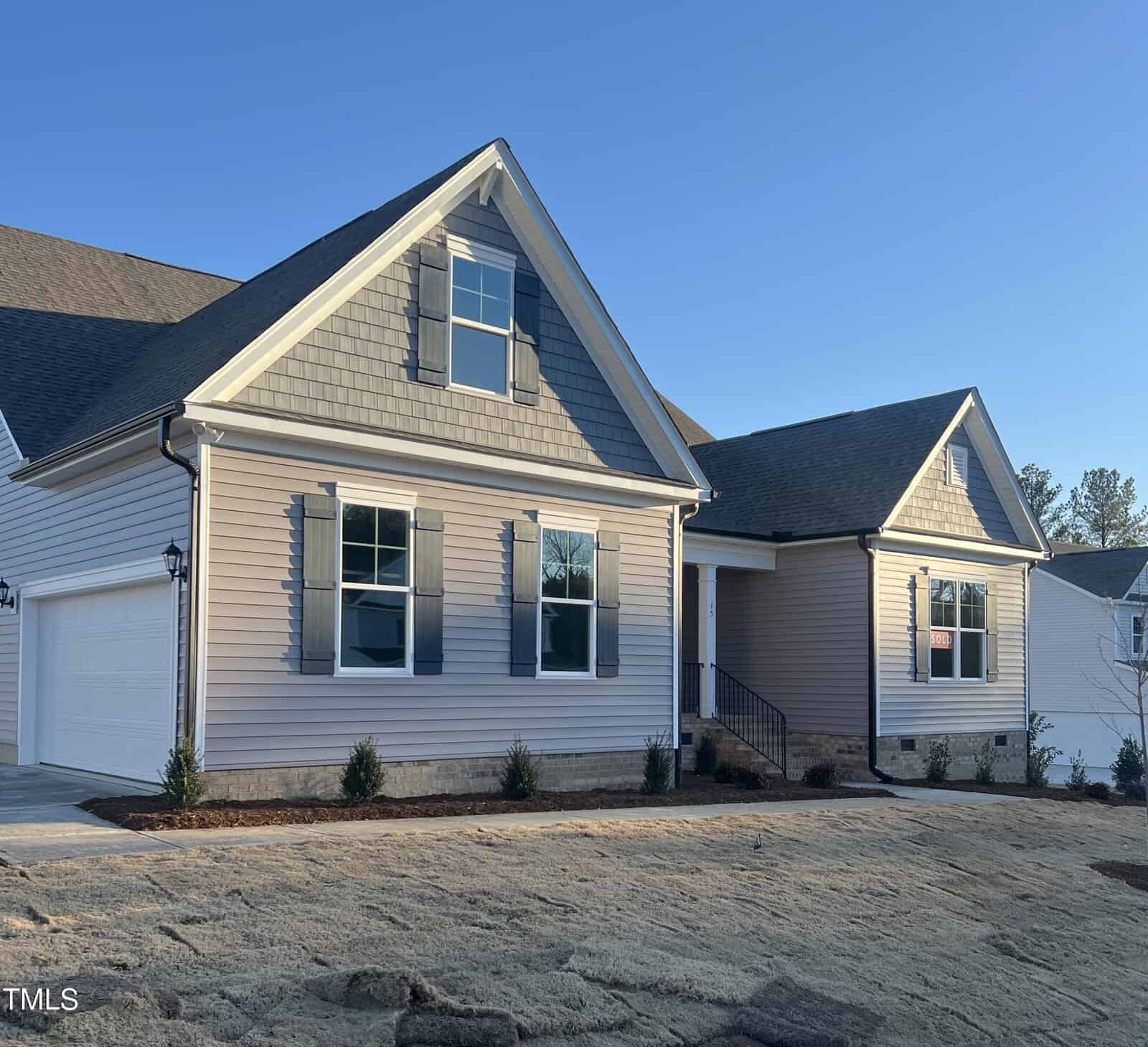 A single-story house with gray siding, white trim, and a two-car garage. The yard has fresh sod and small shrubs planted near the foundation. The sky is clear and blue.