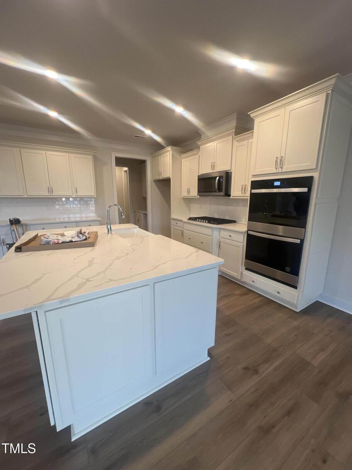 Modern kitchen with white cabinets, marble countertops, double wall ovens, built-in microwave, gas cooktop, stainless steel sink in an island, and wood-look flooring. Bright overhead lighting is visible.