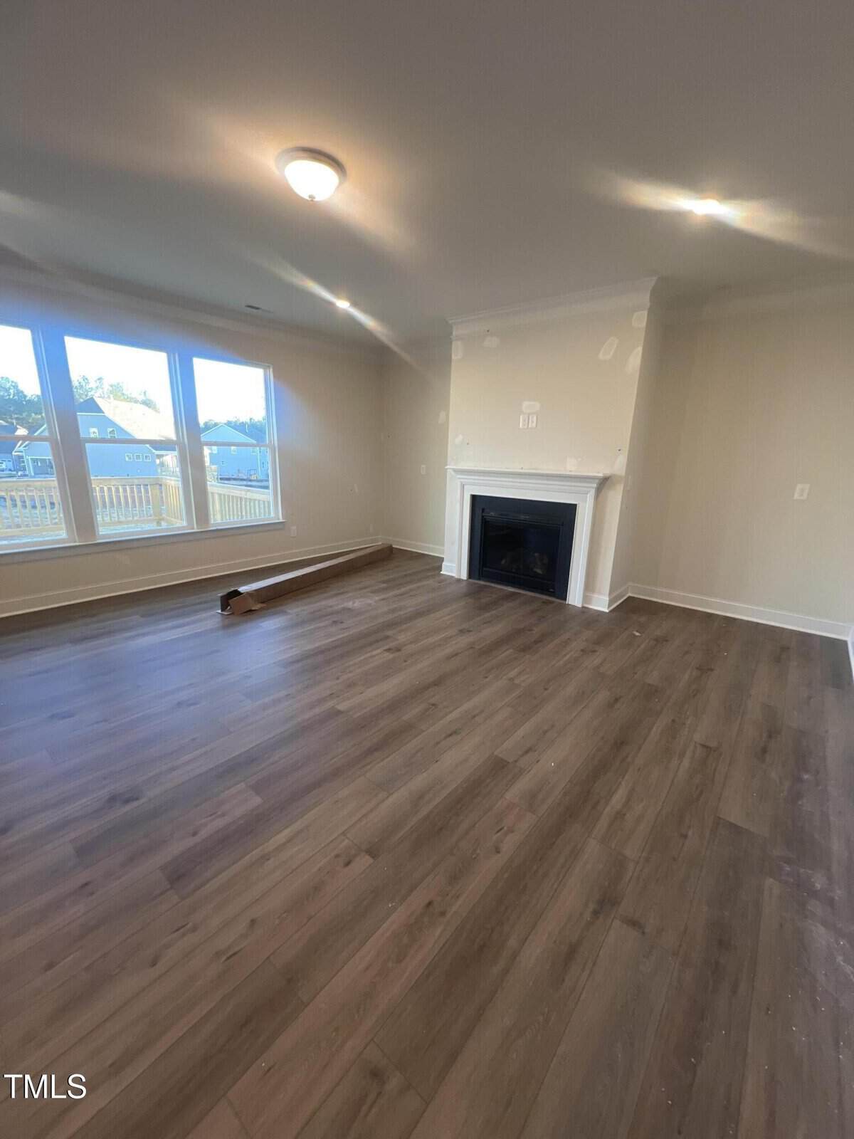 Unfurnished living room with wood flooring, large windows, beige walls, white fireplace, and ceiling light. A rolled-up carpet is near the windows. Sunlight is coming in from the left side.