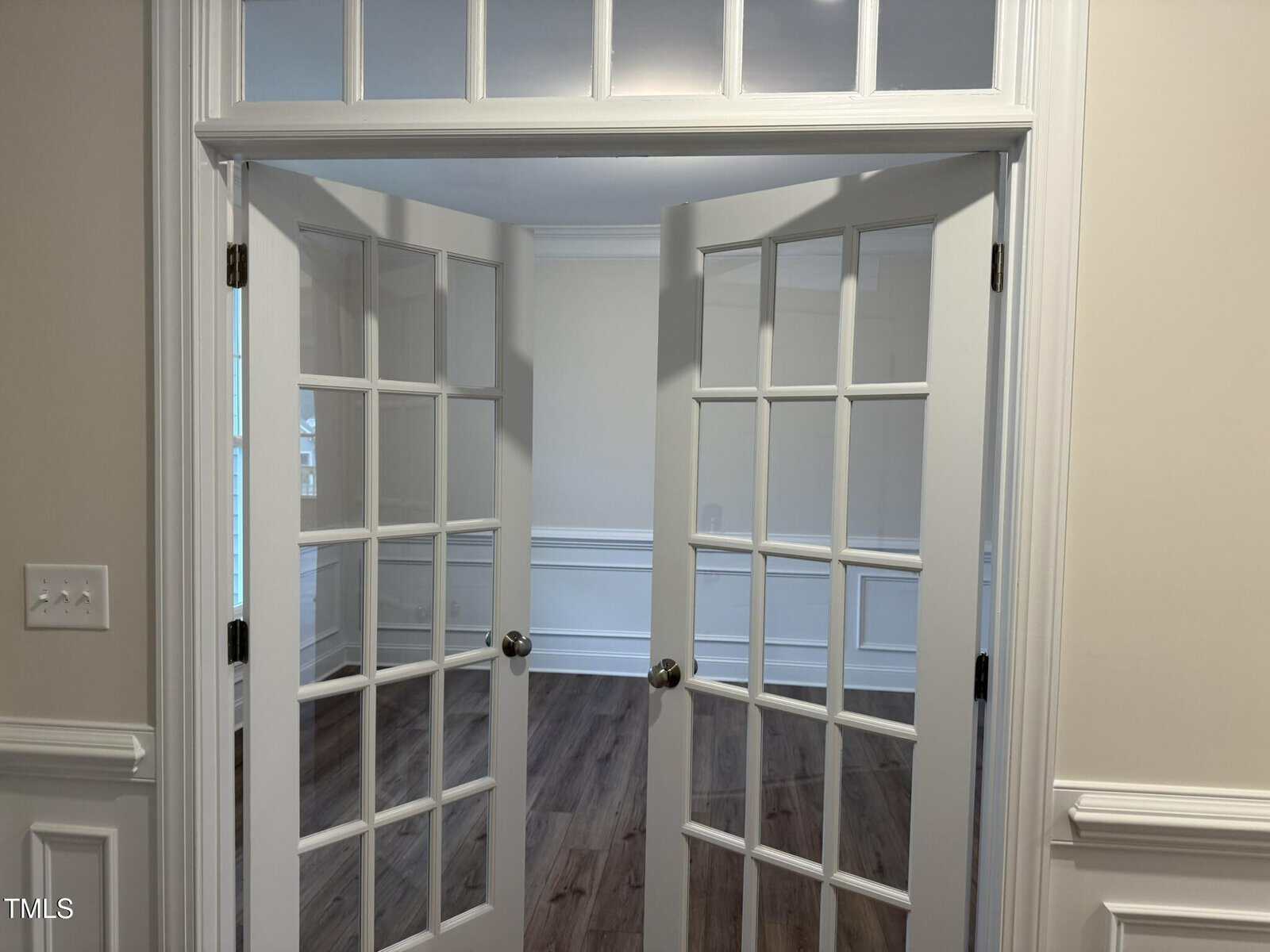A pair of white French doors with glass panes open into a room with white wainscoting, beige walls, and wood-style flooring. The view includes part of a door frame and a light switch on the left wall.