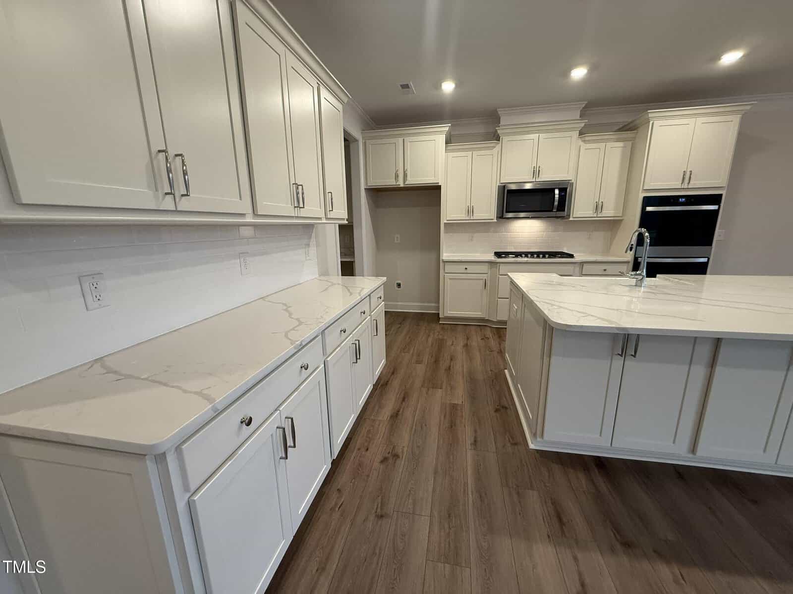 A modern kitchen with white cabinets, marble-look countertops, wood flooring, built-in microwave, oven, gas cooktop, sink on the island, and recessed ceiling lights.