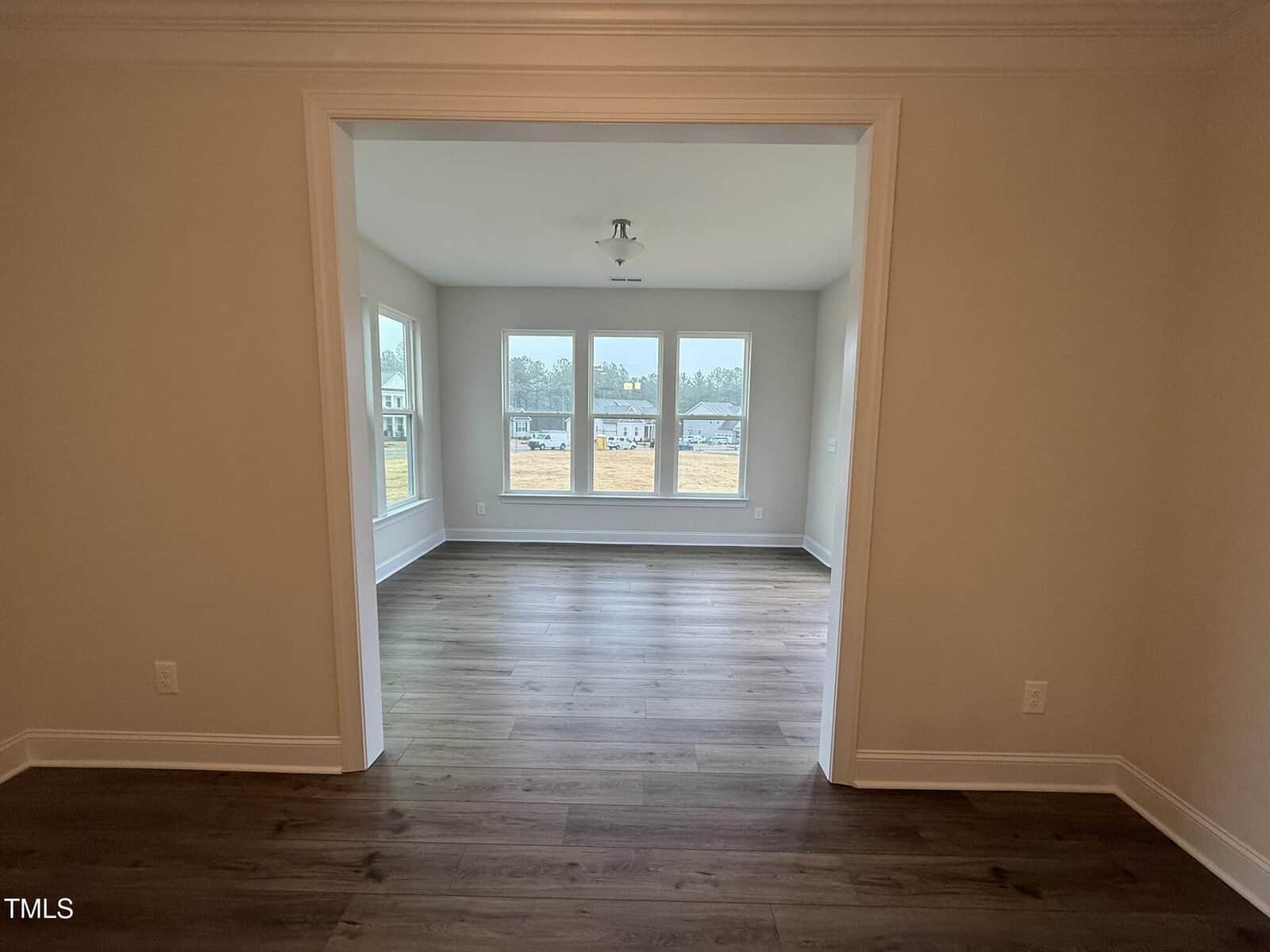 A view from one empty room into another with large windows, wood flooring, beige walls, and a ceiling light fixture. Houses and trees are visible through the windows outside.