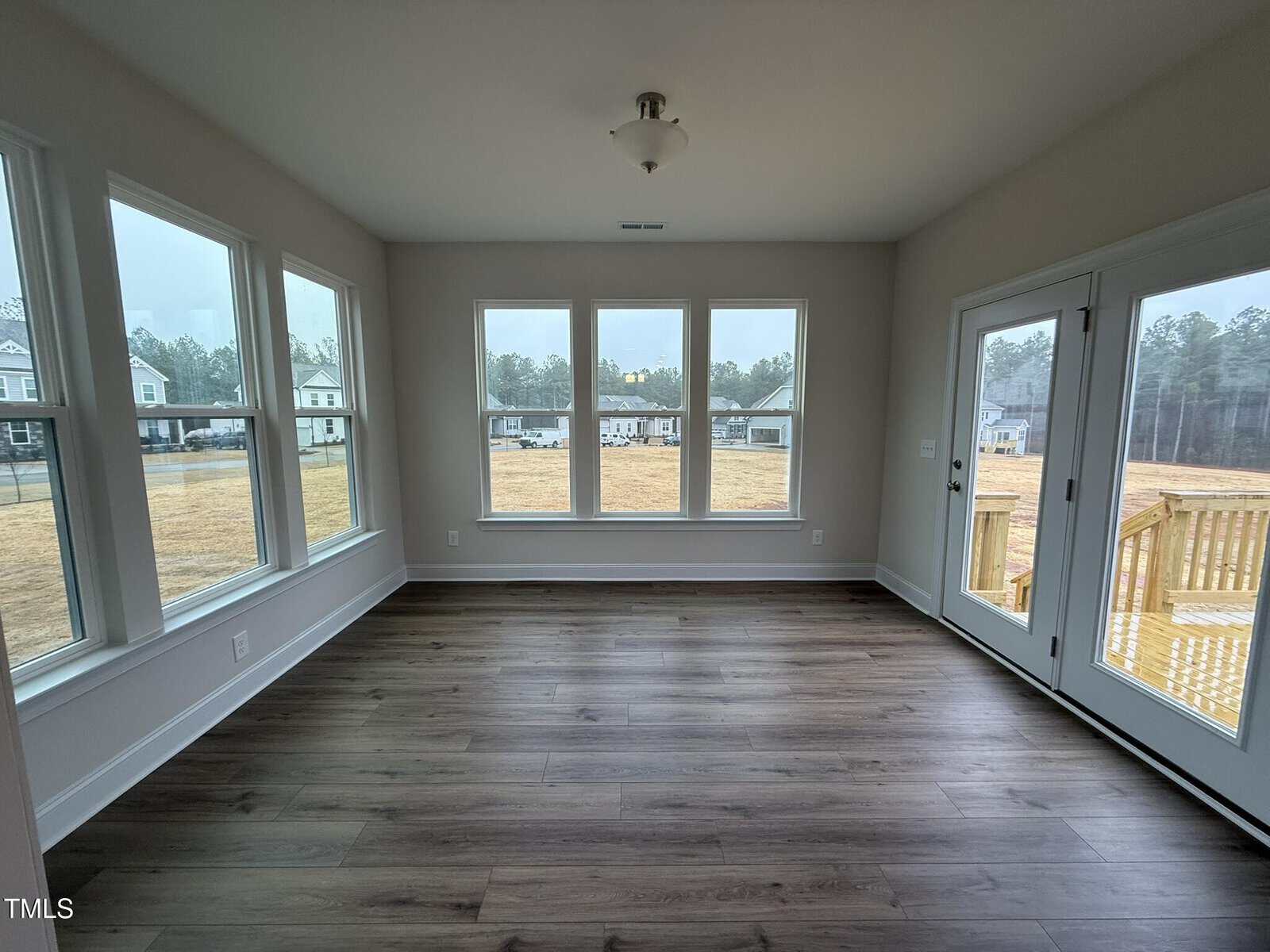 Bright, empty room with light-colored walls, large windows, and double glass doors. Wood-look flooring and ceiling light fixture. Outdoor deck and suburban houses visible through windows.