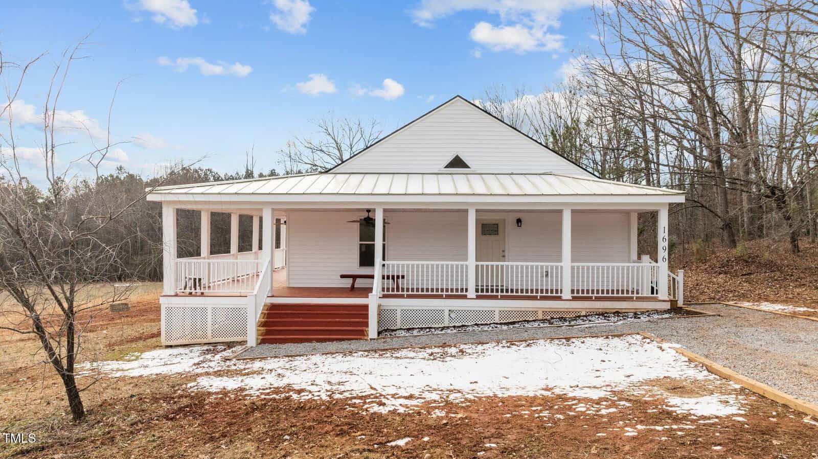A white house on Oxford's 1696 James Winston Road features a metal roof and wrap-around porch, nestled in a rural setting. The ground is dusted with snow, while a gravel path leads to the porch steps. In the background, trees stand under a partly cloudy sky, adding to its serene charm.