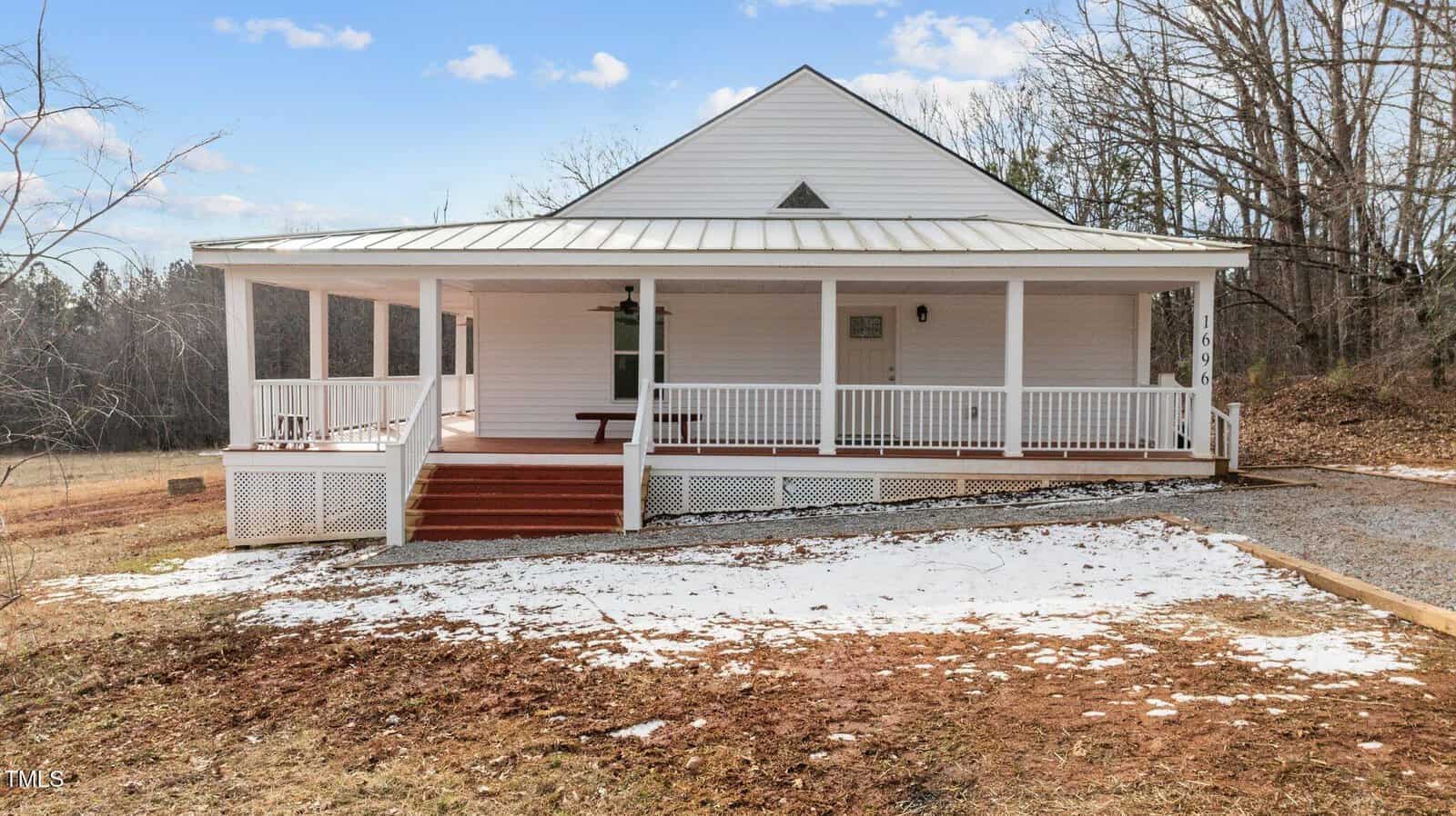 A white, single-story house with a metal roof and a wraparound porch featuring red steps is surrounded by sparse grass and patches of snow. Bare trees and a gravel driveway are seen on a clear day with a few clouds in the sky.