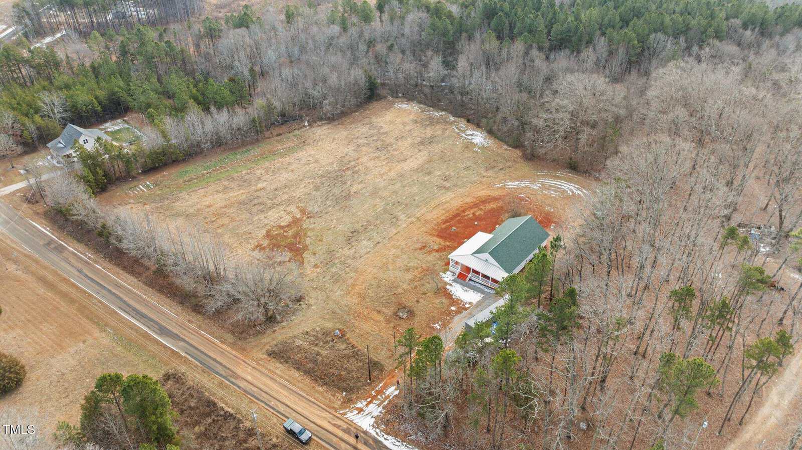An aerial view of a rural landscape featuring a large, open field with patches of brown soil and sparse grass. A house with a green roof is situated on the right. Surrounding the property are trees and a dirt road runs along the left side.