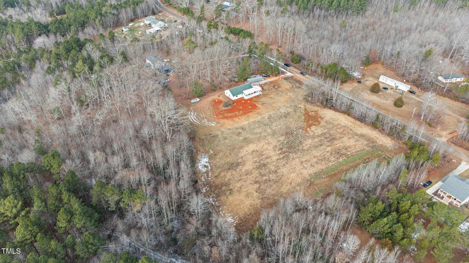 Aerial view of a rural area with sparse tree coverage. Central buildings include a white house and barn surrounded by red earth. Dirt paths and a few scattered houses are visible. The surroundings include grass fields and leafless trees.
