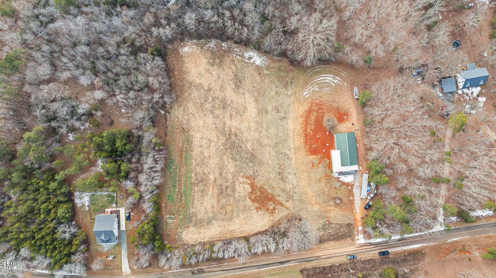 Aerial view of a rectangular, grassy field surrounded by trees, with two buildings at the edge. There is a dirt area next to the larger building, and a road borders the field at the bottom of the image.