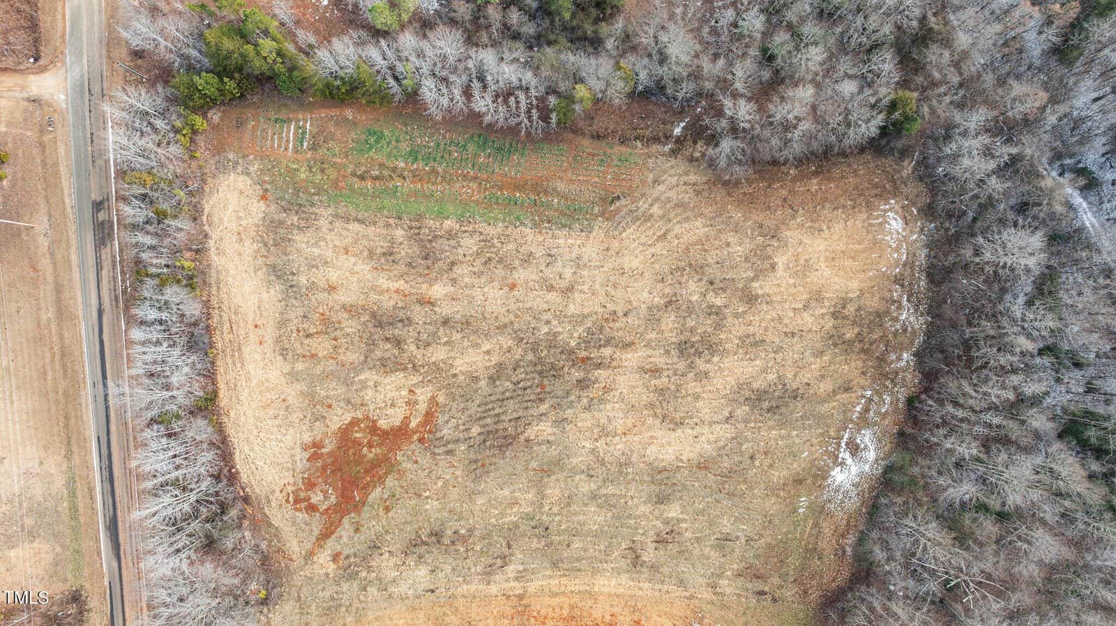 Aerial view of a barren field bordered by trees and a road on one side. The field has patches of green and brown, with a tree line surrounding most of its perimeter. Sparse vegetation and visible soil erosion are present.