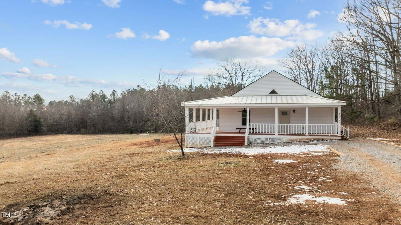 A small, white, single-story house with a wraparound porch stands in an open field with sparse patches of snow. Leafless trees border the area, and a gravel path leads to the front steps. The sky is partly cloudy.