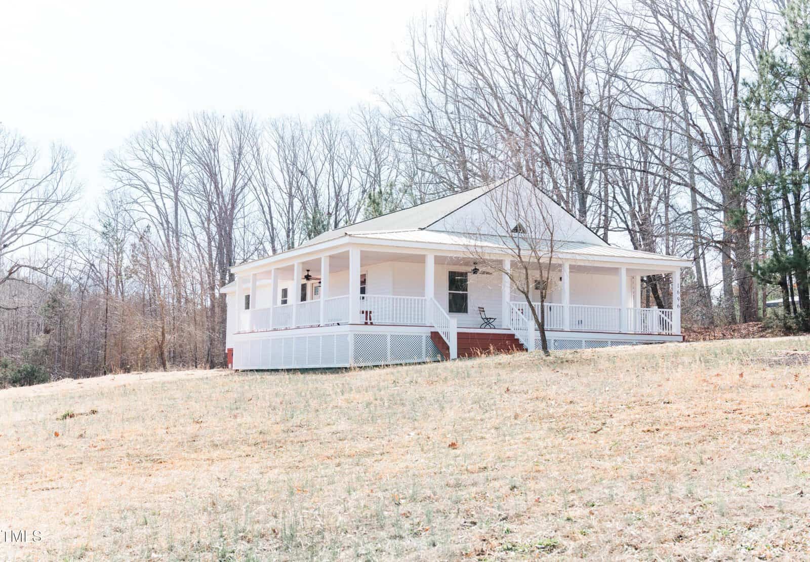 A white, single-story house with a covered front porch is situated on a grassy hill. Leafless trees and evergreens surround the house, and a single chair is visible on the porch. The sky is clear.