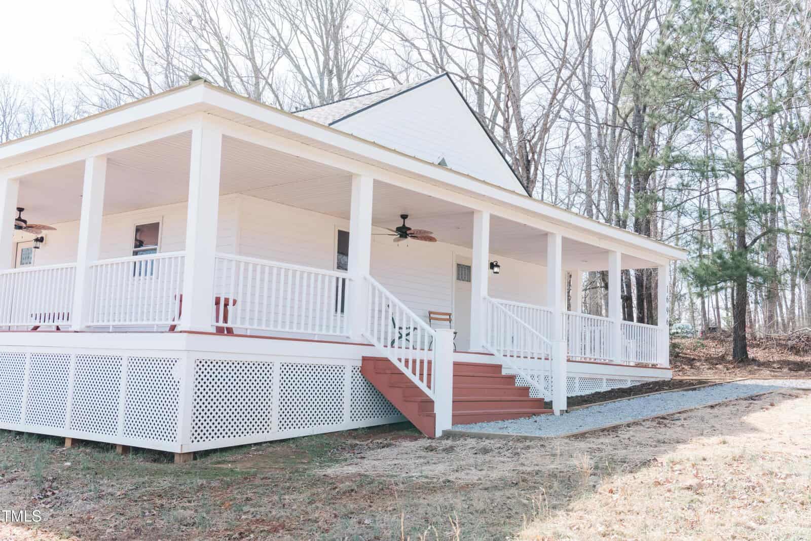 A white house with a wide front porch featuring railings and steps leading up to the entrance. Ceiling fans are installed on the porch. The house is set against a backdrop of leafless trees and a small grassy area with a gravel path.