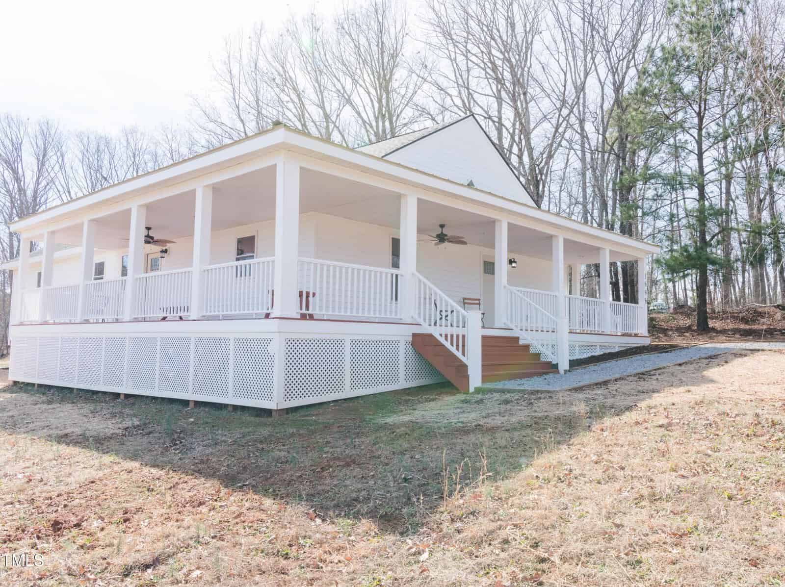 Single-story white house with a wraparound porch, situated on a grassy plot. The porch is supported by multiple white columns and features ceiling fans. Bare trees and some evergreens are in the background. Steps lead up to the porch at the front.
