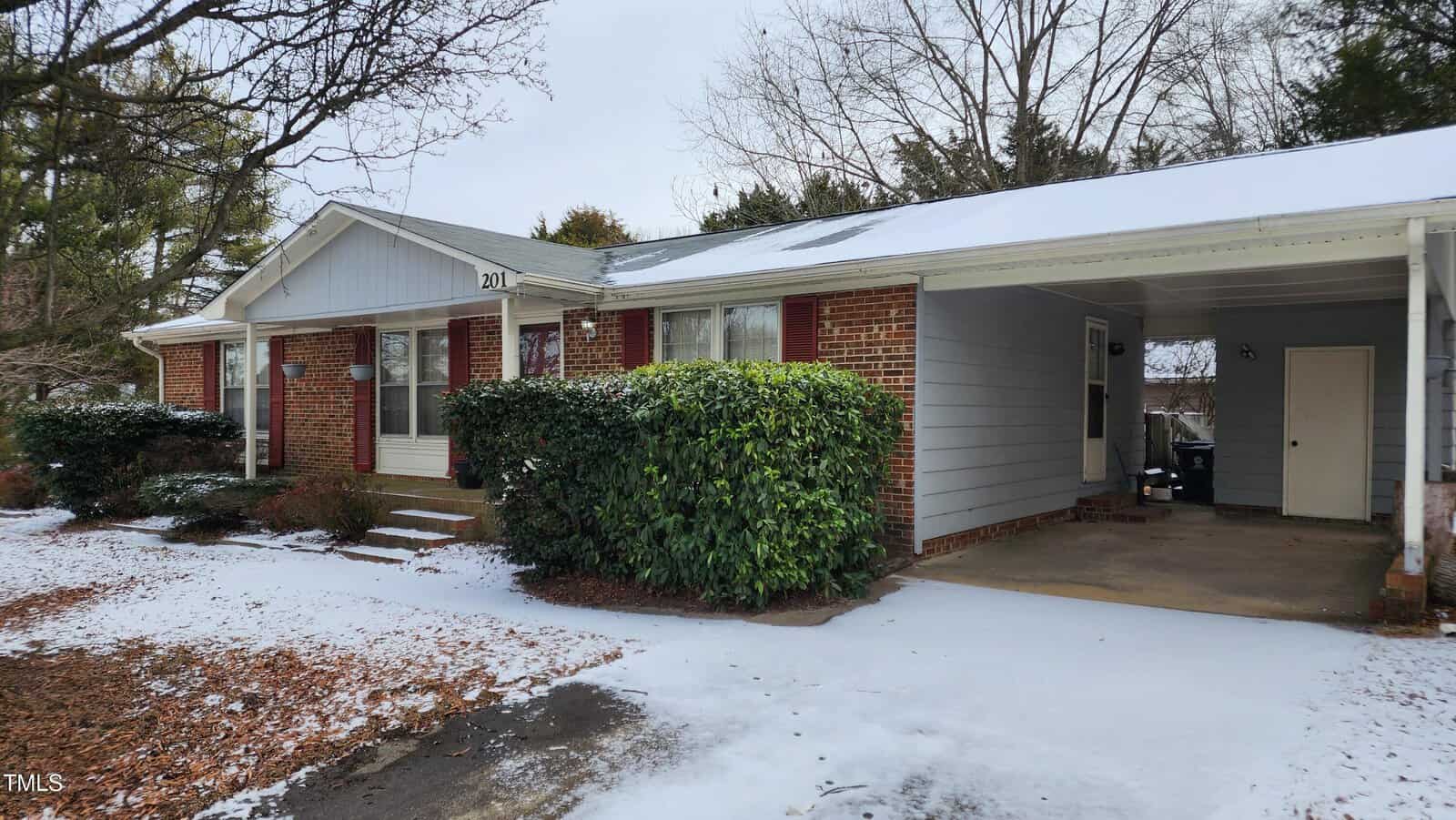 Single-story brick house with carport, surrounded by light snow. The front features steps leading to a door, flanked by windows with shutters, and trimmed bushes. Leafless trees and some evergreens are visible in the background.