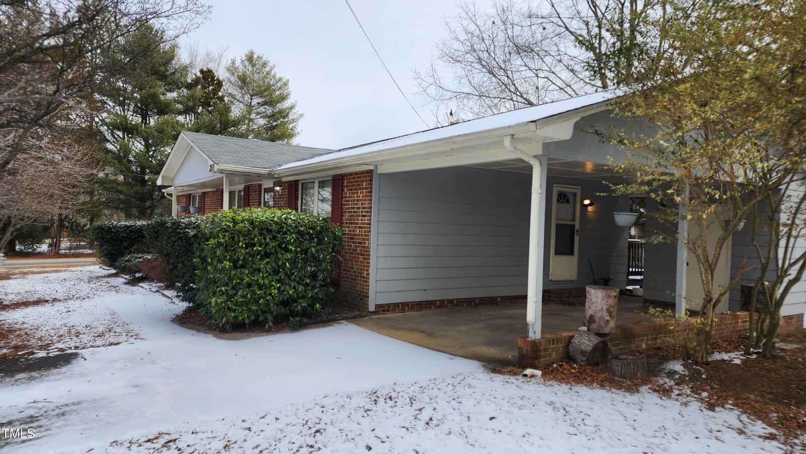 A single-story brick and siding house with a covered entryway is surrounded by snowy ground. Bushes line the front, and deciduous trees are visible in the background. The roof is greenish, and there is a small pile of chopped wood near the doorway.