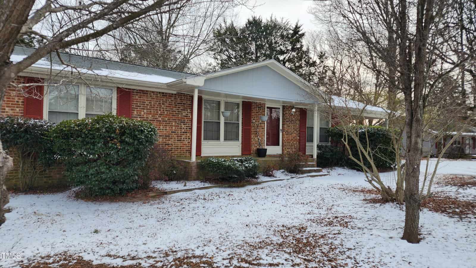 A single-story brick house with a snow-covered yard. There are red shutters, bushes near the windows, and bare trees in the front yard. The sky is overcast.