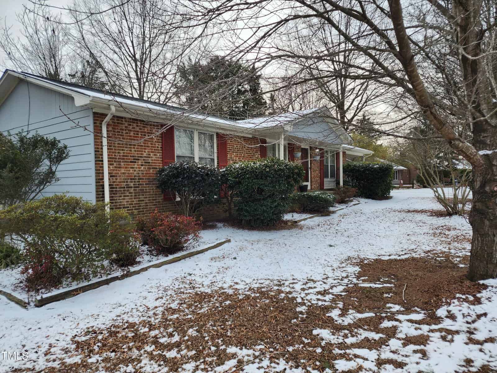 A single-story house with a brick and gray facade is surrounded by leafless trees and bushes. A light layer of snow covers the ground, creating a wintry scene. The house has red shutters and a neatly trimmed yard.