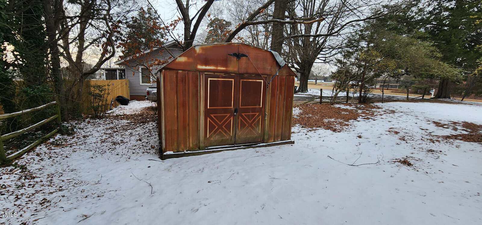 A small, rust-colored shed with peeling paint stands in a snow-dusted yard. It has double doors with diagonal wooden accents. The background includes a house, trees with bare branches, and a lightly snow-covered ground.