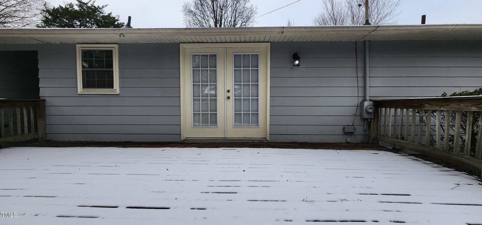 A snow-covered wooden deck leads to a light gray house with a set of French doors in the center. The doors are flanked by a window on the left and a black outdoor light on the right. Trees are visible in the background.