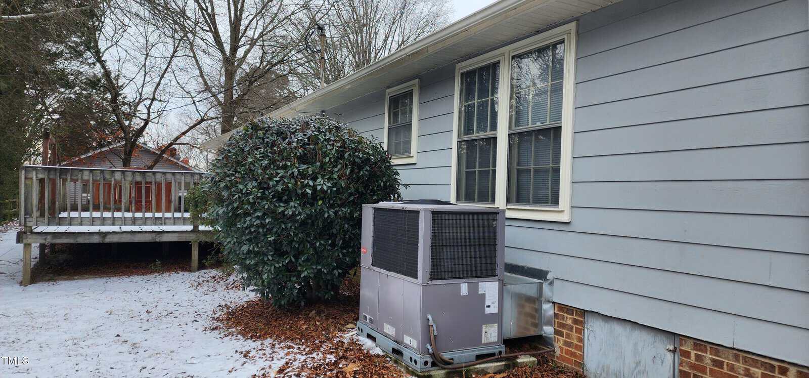 A gray building with a three-pane window is shown. An air conditioning unit sits on the ground beside the building. Snow and fallen leaves cover the ground, with a large shrub nearby. A wooden deck is visible in the background.