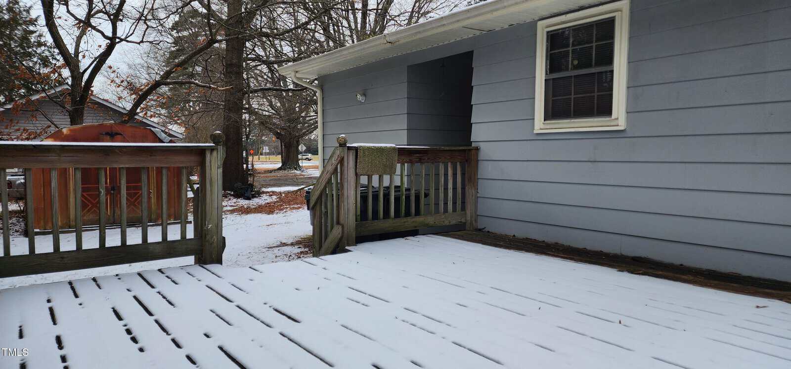 A wooden deck and steps covered in a light layer of snow are attached to a gray house. Trees with bare branches are visible in the background, and a brown shed is partially seen to the left. The scene is overcast and wintry.