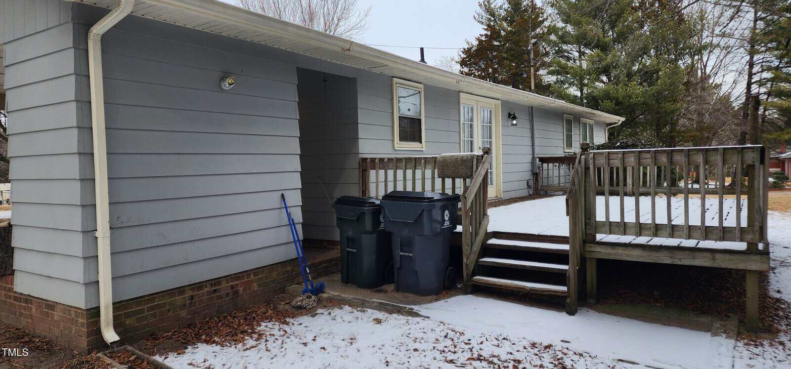 A small, single-story house with light gray siding and a brick foundation. A wooden deck with stairs leads to the rear door. Two trash bins and a snow-covered ground are visible. Several trees stand in the background.