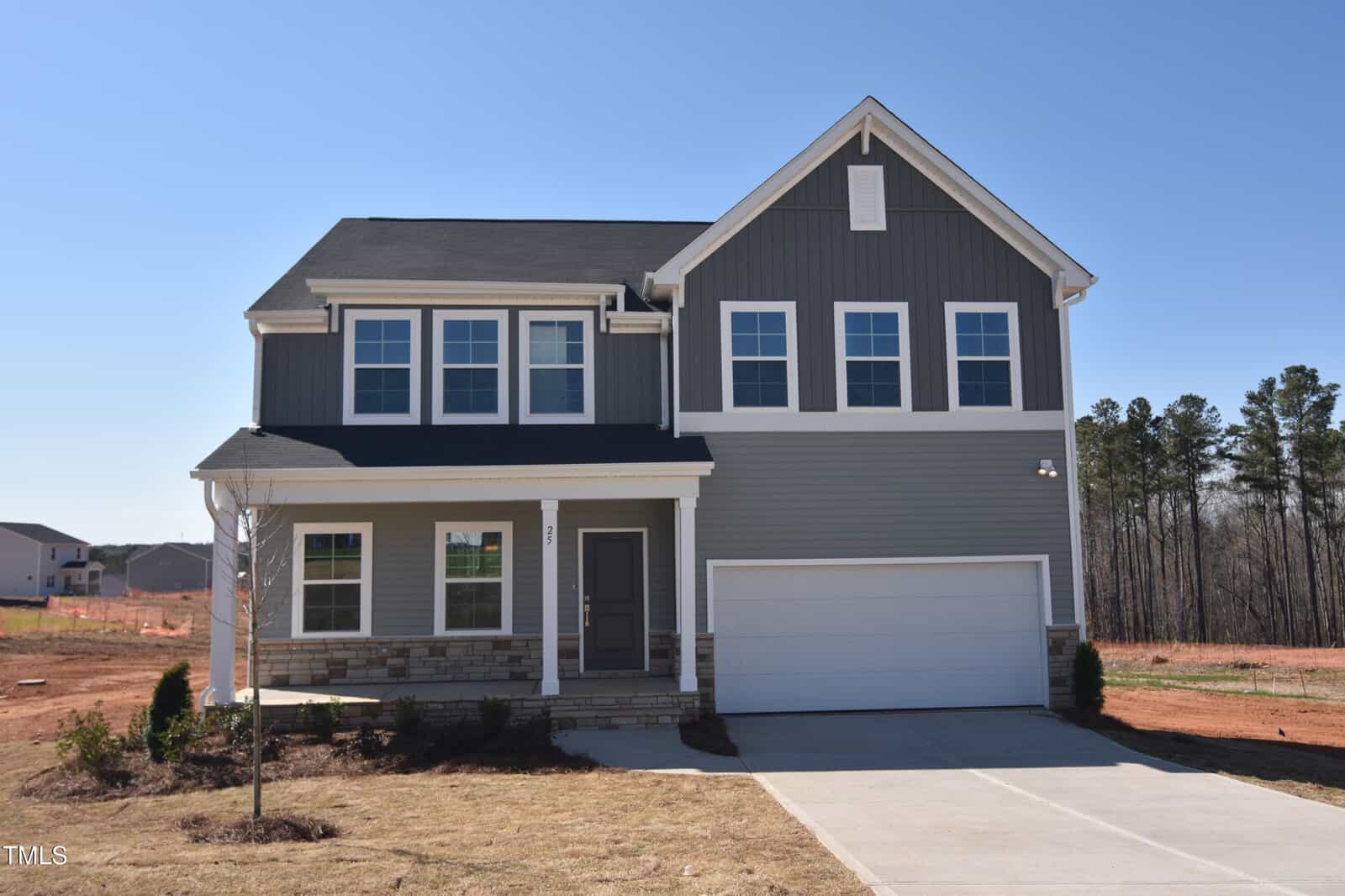 Two-story modern house with gray siding, white trim, and stonework at the base. It has a two-car garage, a small covered front porch, and minimal landscaping. The sky is clear and the lot is newly developed.