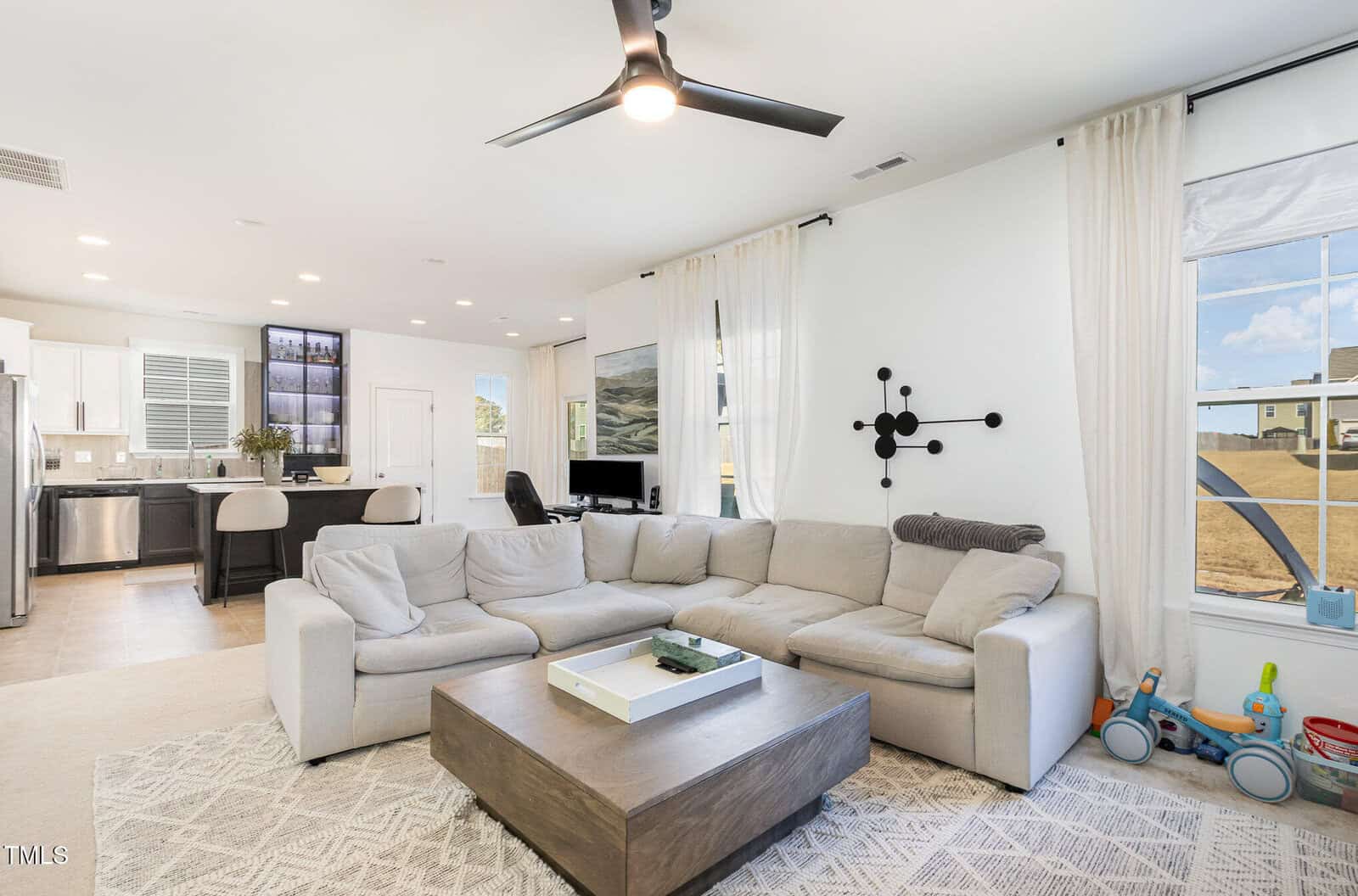 A modern living room with a large white sectional sofa, square wooden coffee table, wall clock, ceiling fan, and open view to a kitchen with barstools. Large windows with white curtains let in natural light.