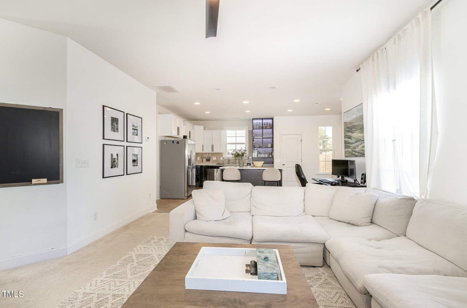 A modern living room with a white sectional sofa, a coffee table, wall art, a TV, and an open view to a kitchen with bar stools and stainless steel appliances. Large windows let in natural light.