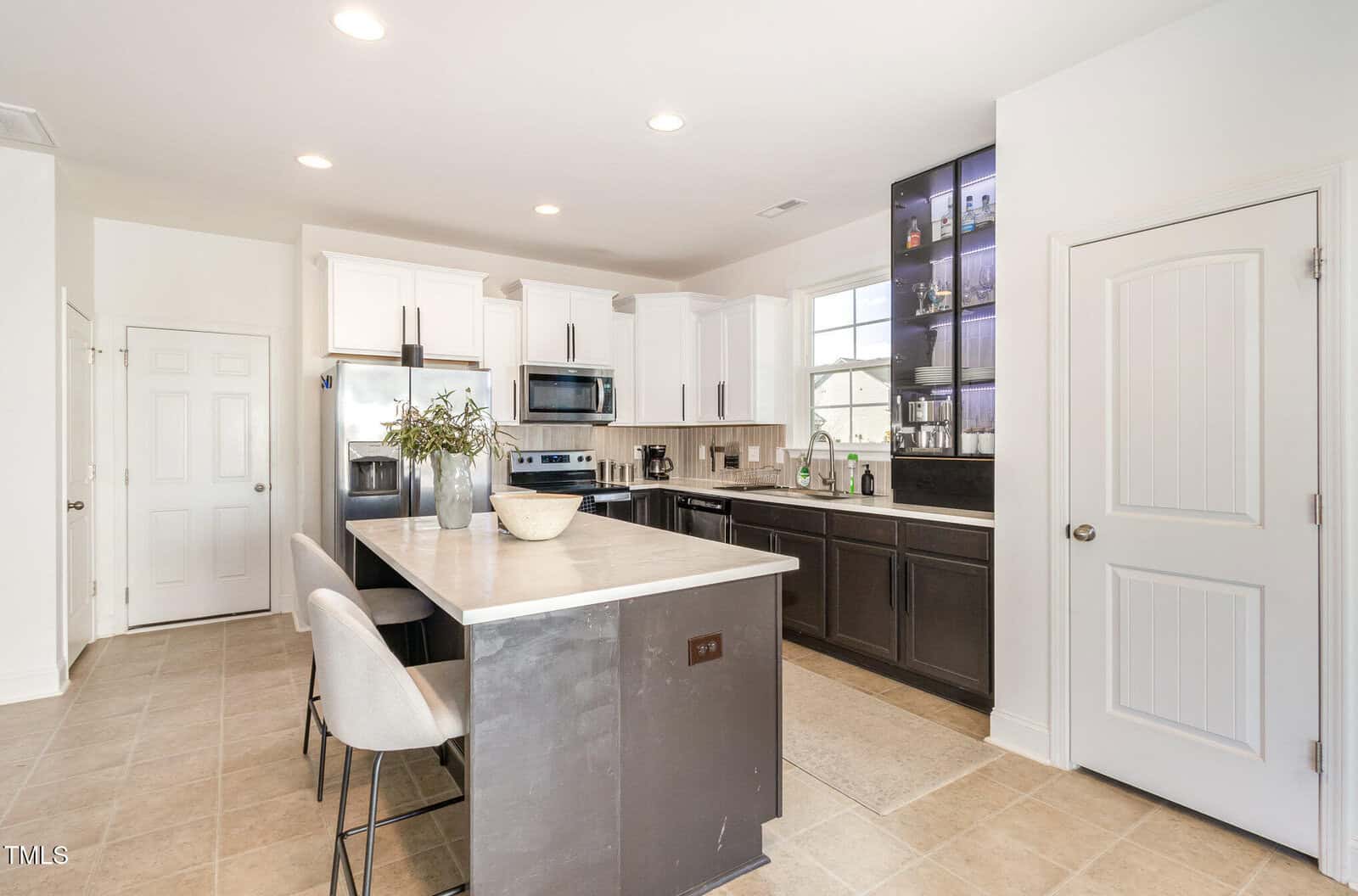 Modern kitchen with white upper cabinets, dark lower cabinets, stainless steel appliances, a center island with two barstools, a vase with greenery, tiled floor, and a door on the right. Shelves with glassware are backlit.