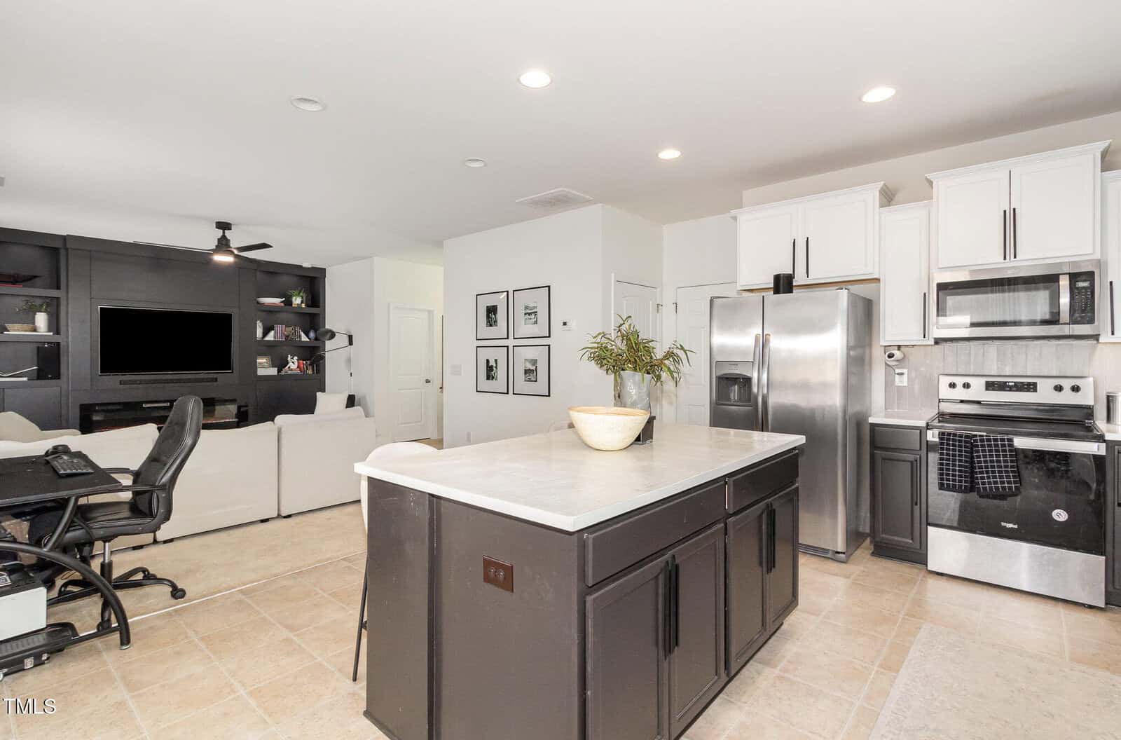 Modern kitchen with a large island in the center, stainless steel appliances, white upper cabinets, and black lower cabinets. Open layout leads to a living area with a TV, couch, wall art, and a desk workspace.