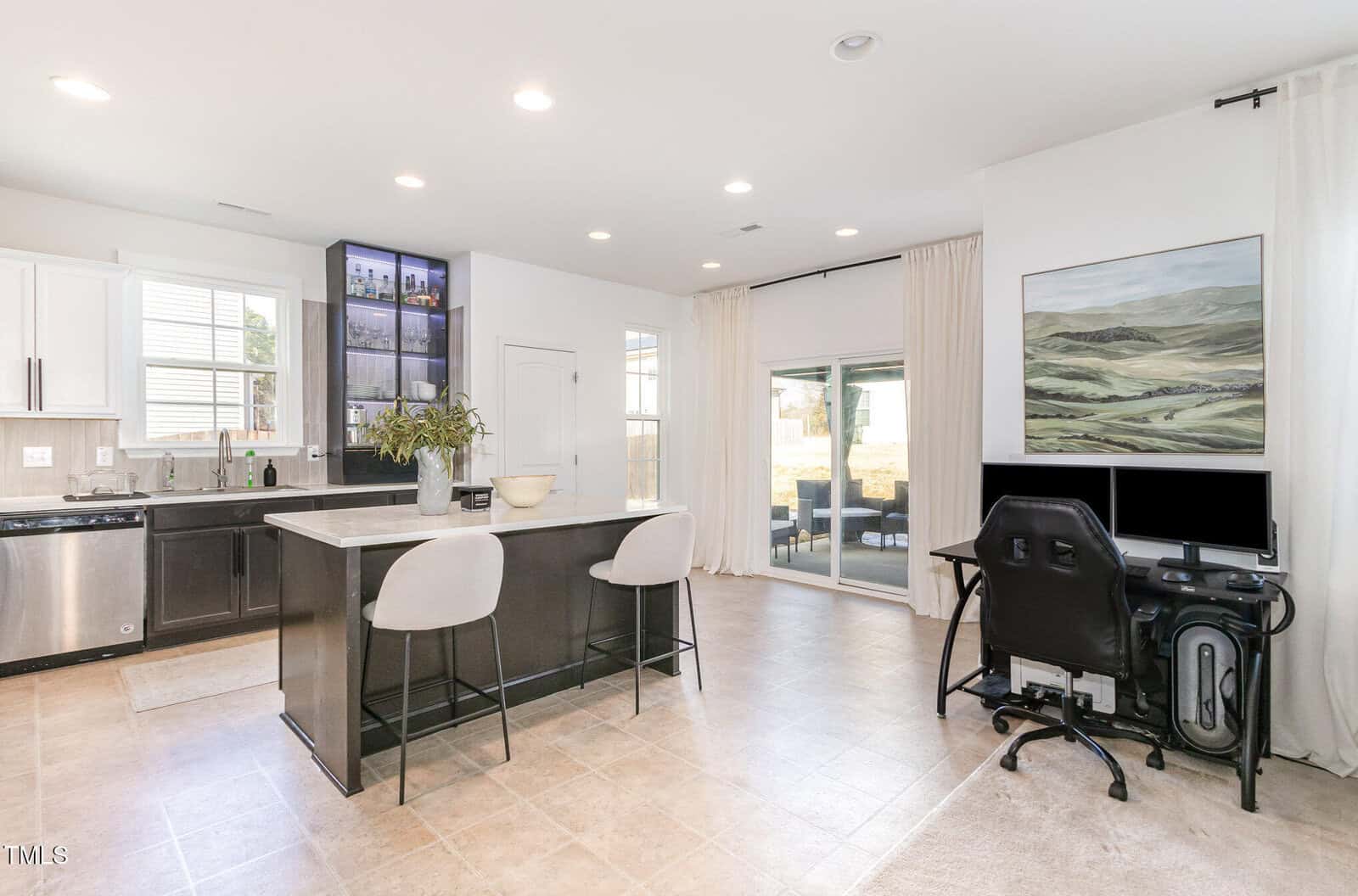 Modern kitchen and living space with a black island and two chairs, white cabinetry, a computer desk with dual monitors, sliding glass doors, and a large landscape painting on the wall.