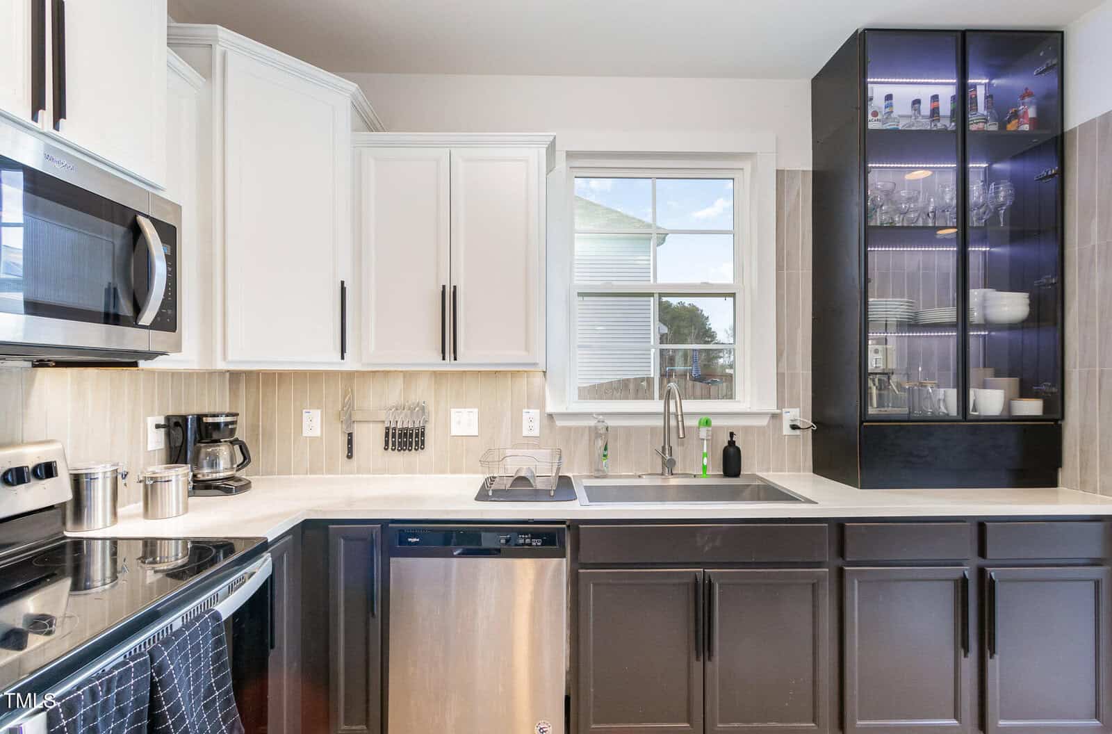Modern kitchen with white upper cabinets, dark lower cabinets, stainless steel appliances, a window above the sink, beige backsplash, and a glass-front cabinet displaying glassware and dishes on the right side.