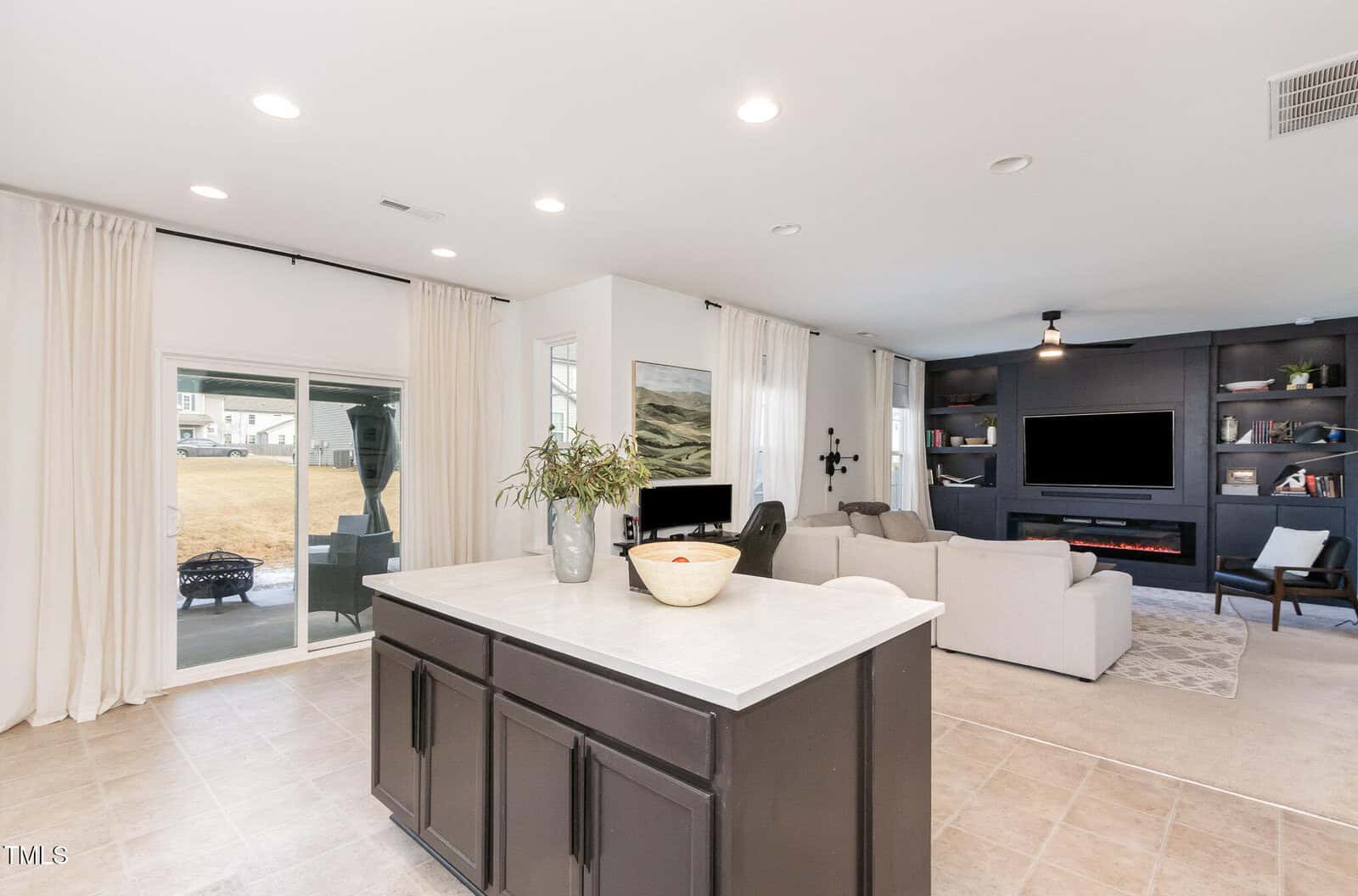 A modern open-concept living space with a kitchen island in the foreground, light tile flooring, white walls, built-in shelving, and large windows with white curtains overlooking a backyard.