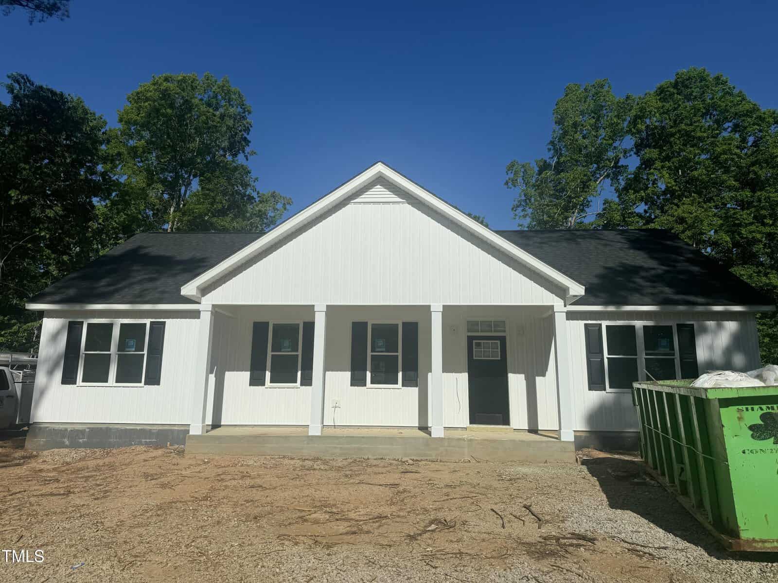 A newly constructed single-story white house with black roof shingles, a covered front porch, and an unfinished yard. A green dumpster sits to the right, and trees are visible in the background under a clear blue sky.