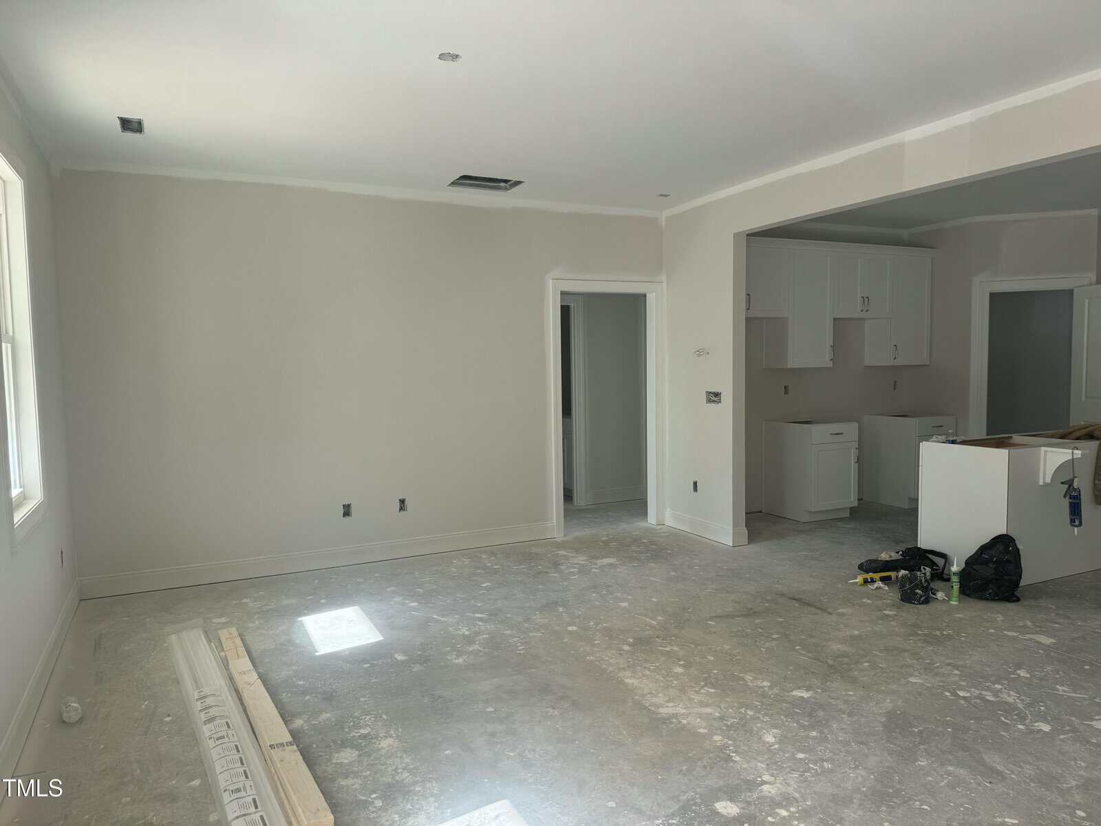 Unfinished room with bare concrete floors and white walls. Some tools and construction materials are on the floor. The kitchen area in the background has white cabinets and an unfinished island.