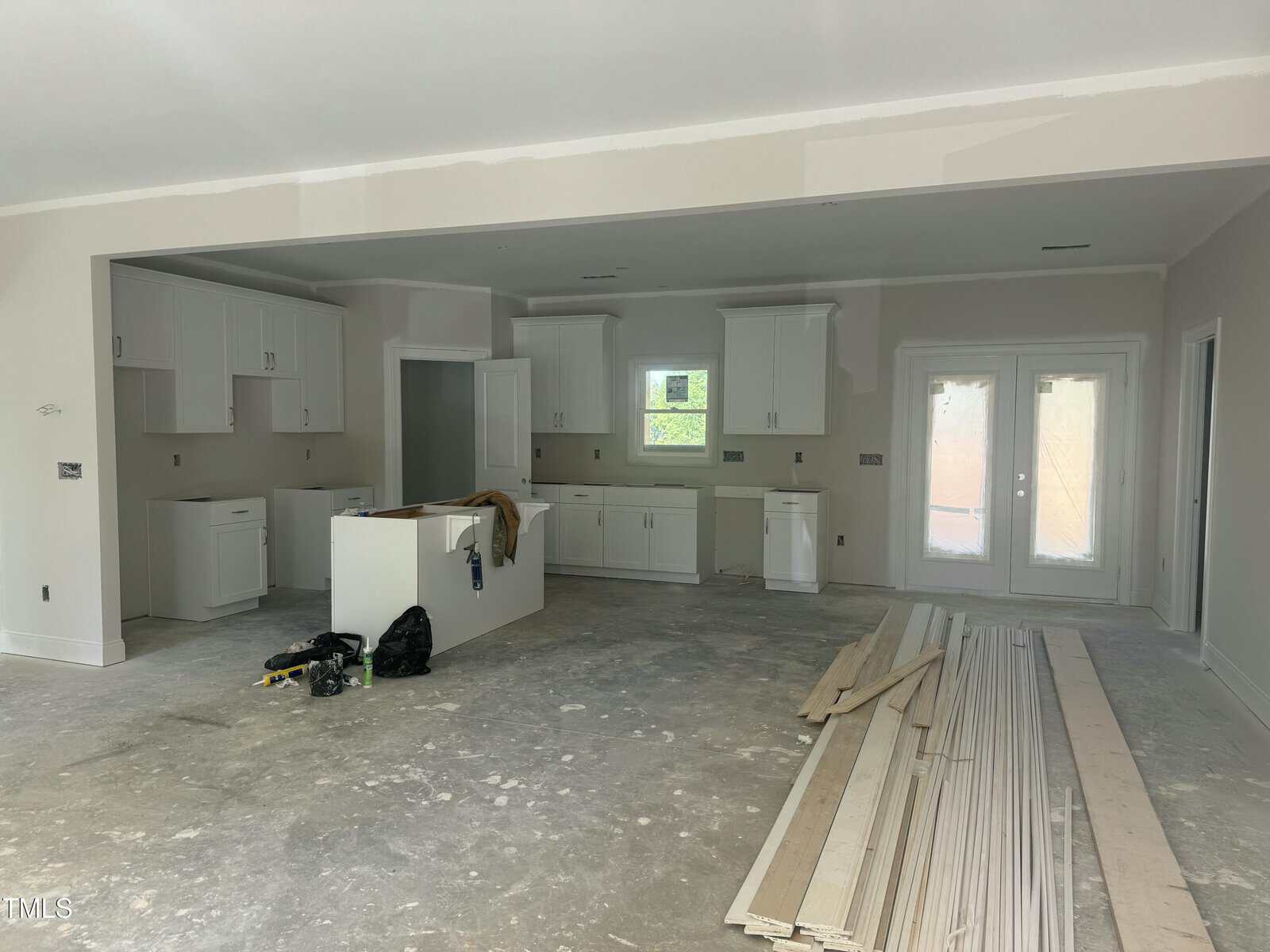 A partially finished kitchen with white cabinets, an unfinished floor, construction materials and tools scattered around, and a few wooden planks stacked in the foreground. French doors and windows are visible in the background.