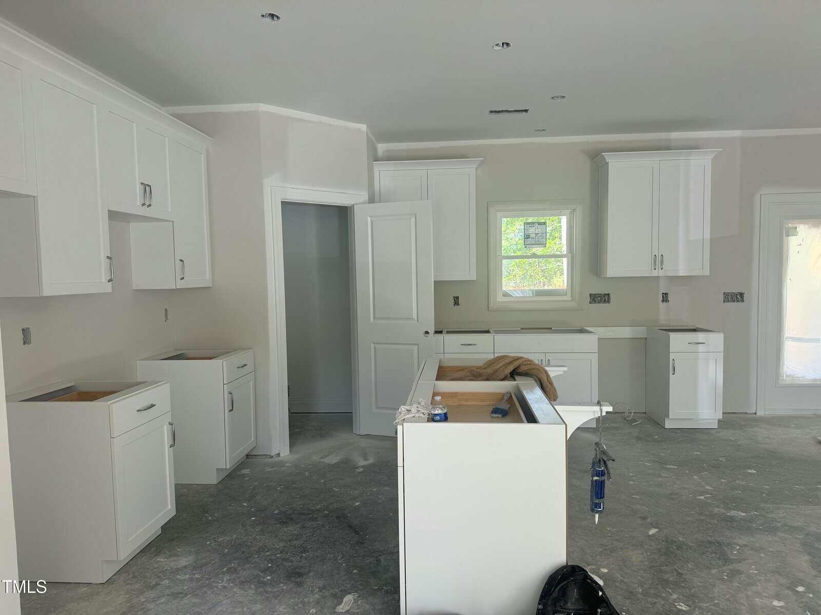 A kitchen under construction with white cabinets and drawers installed, unfinished countertops, open cabinet doors, a window above the sink, construction materials on the floor, and exposed drywall.