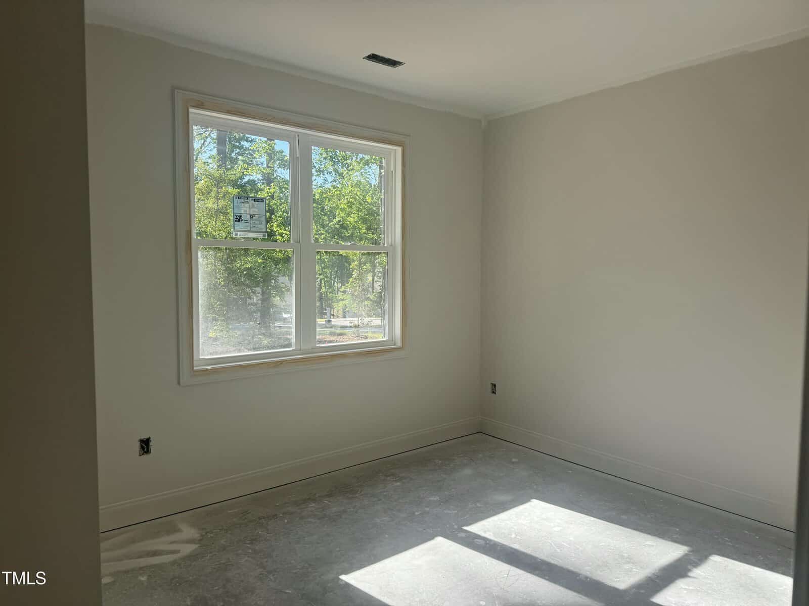 Empty room with unfinished floors, off-white walls, and a large double-hung window letting in sunlight. The window displays a sticker, and the room appears to be under construction or renovation.