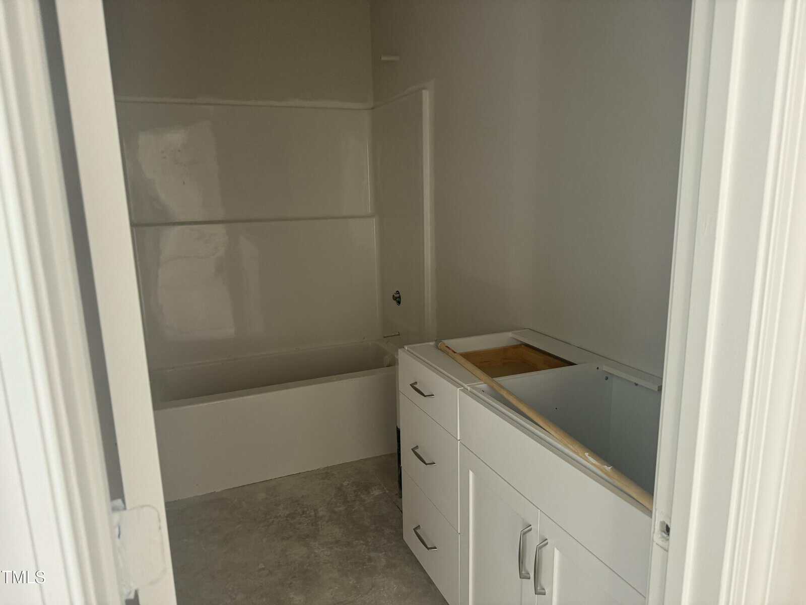Unfinished bathroom with a white bathtub-shower combo and a white vanity with no countertop or sink, set on a bare concrete floor. Walls are plain and unpainted. No fixtures are installed.